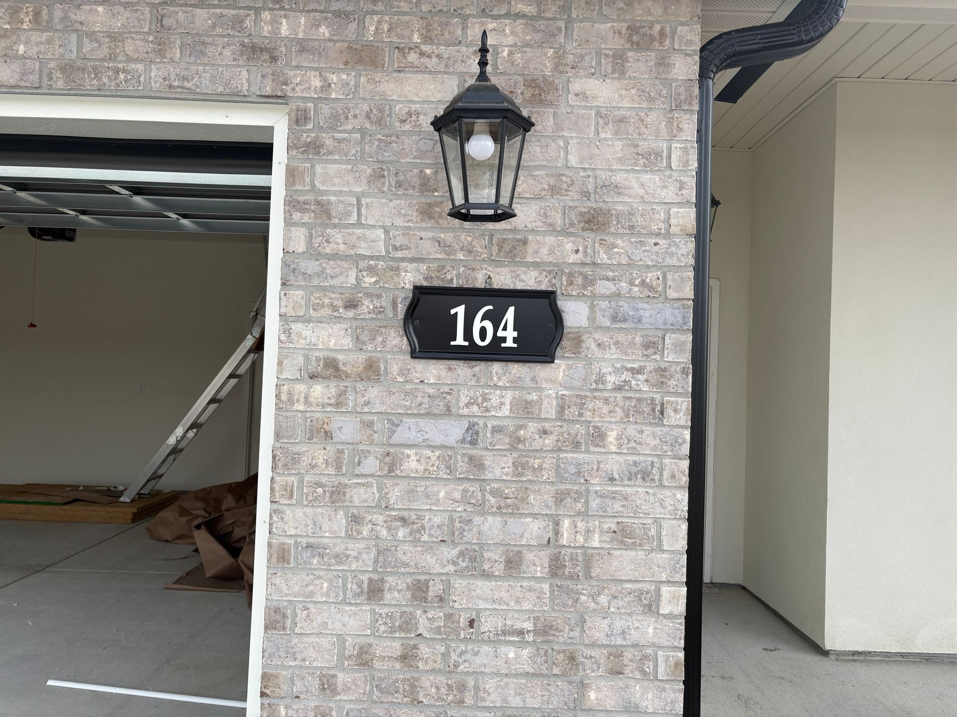 House number 164 on a black plaque, mounted on a tan brick wall beneath a black outdoor wall lamp.