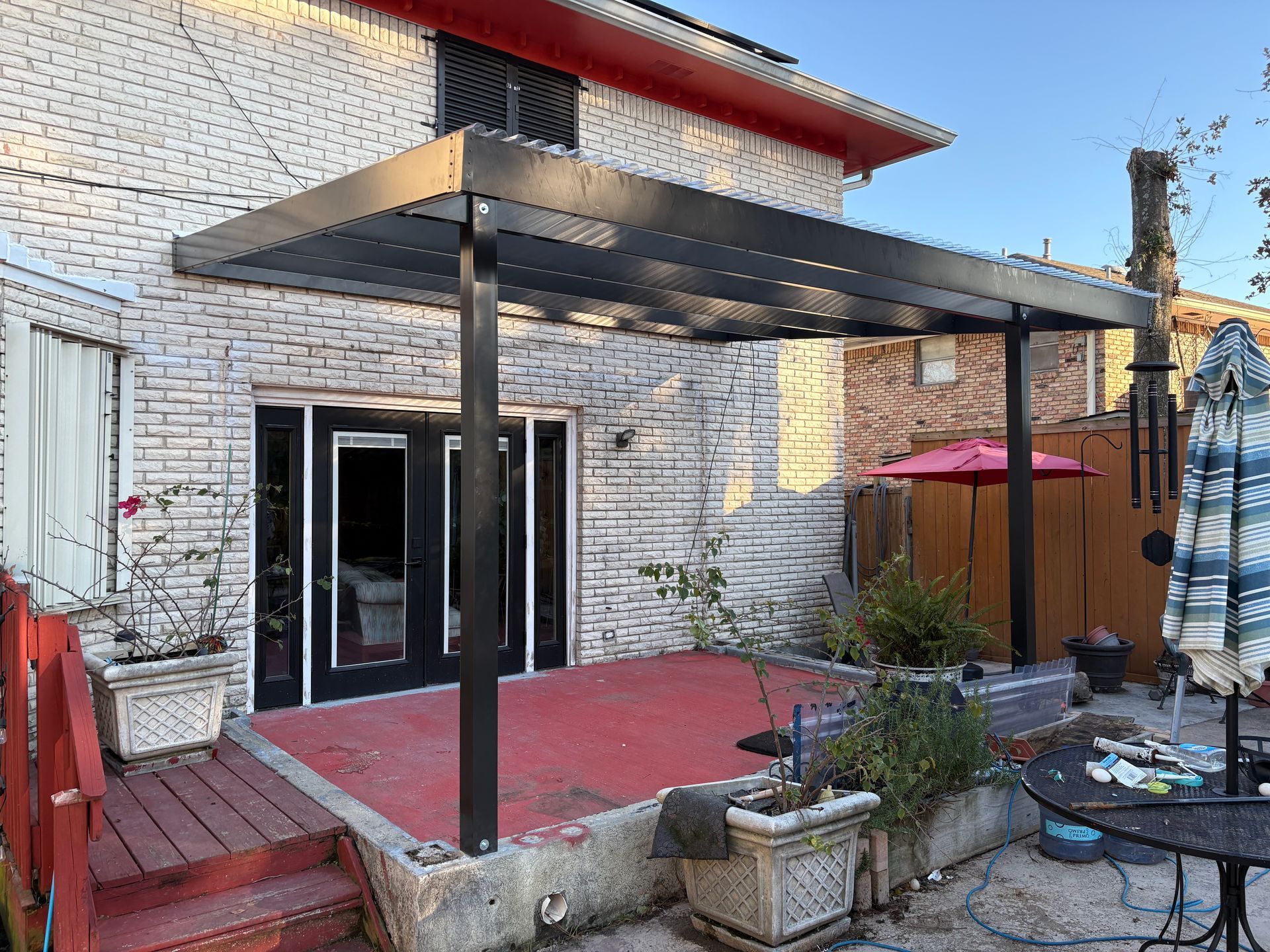 A black pergola stands over a red patio outside a brick house with black-trimmed French doors.