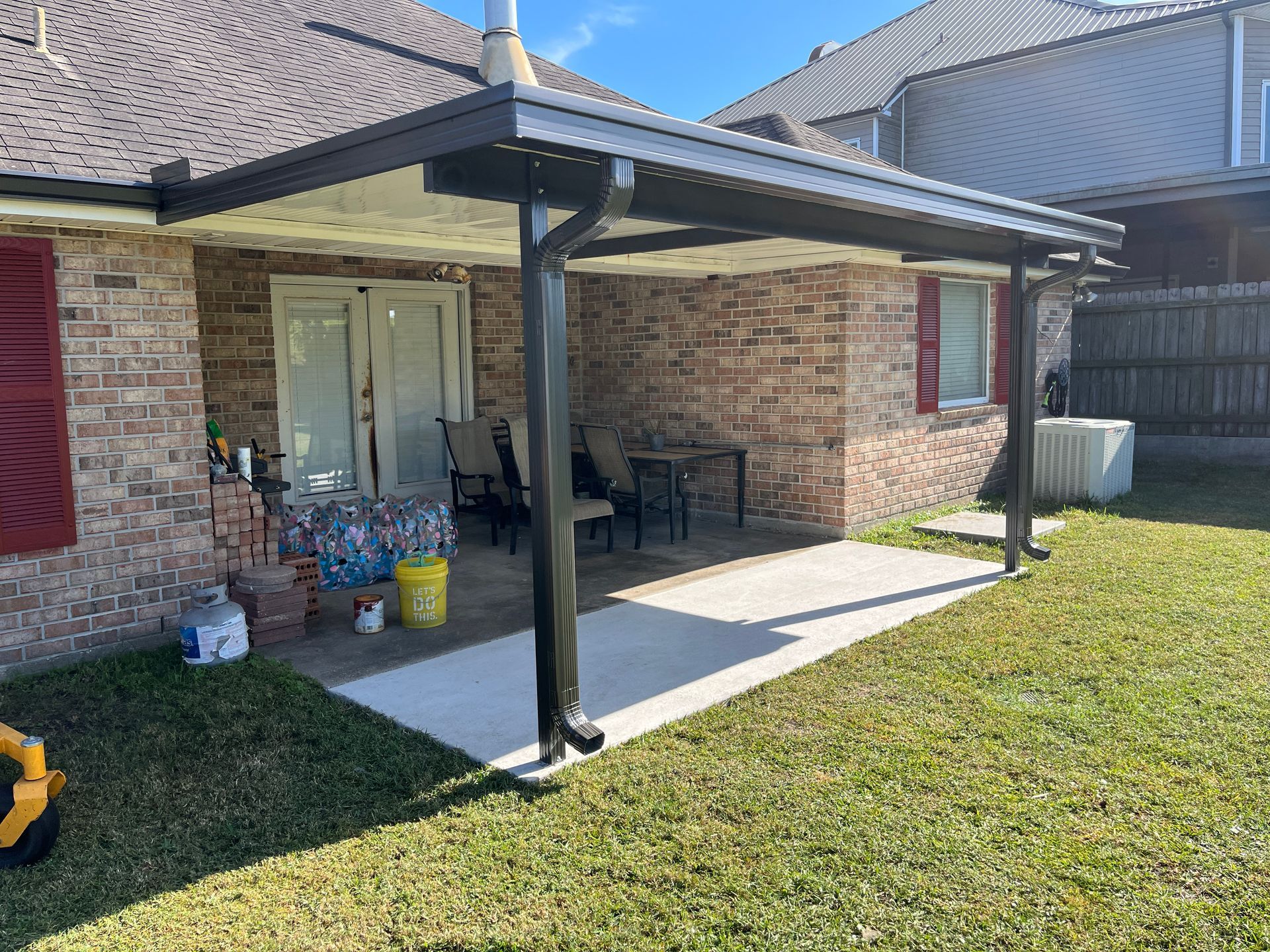 A brick house patio features a covered roof supported by a black column with an attached downspout and outdoor furniture.