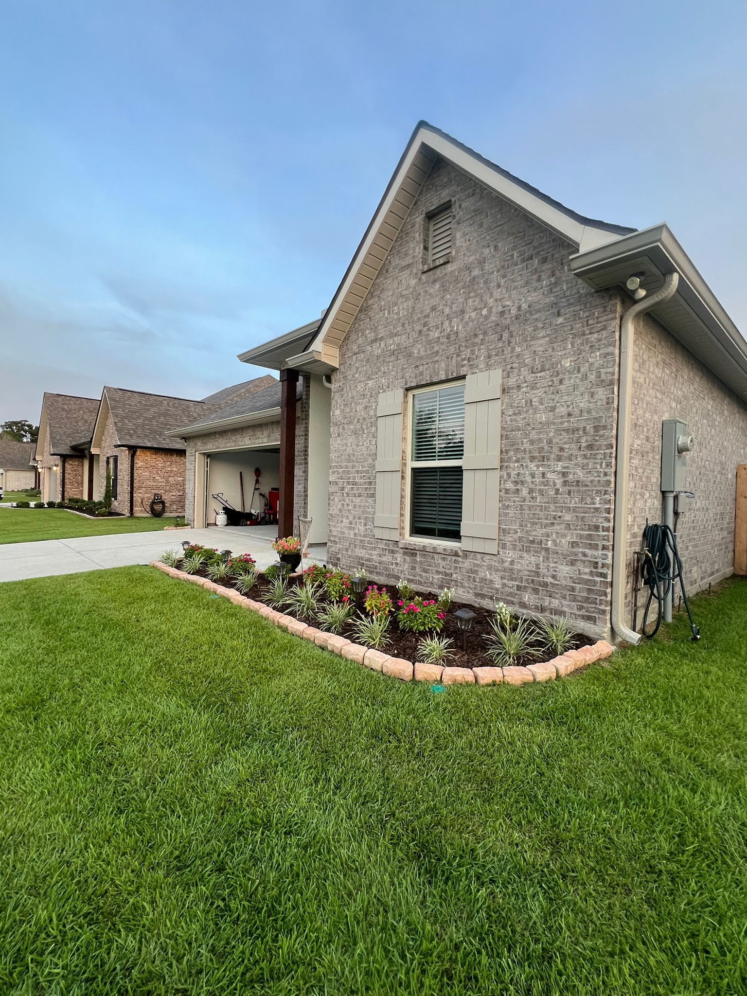 A brick home with a gray shingled roof, a front lawn, and a curved garden bed with small plants under a blue sky.