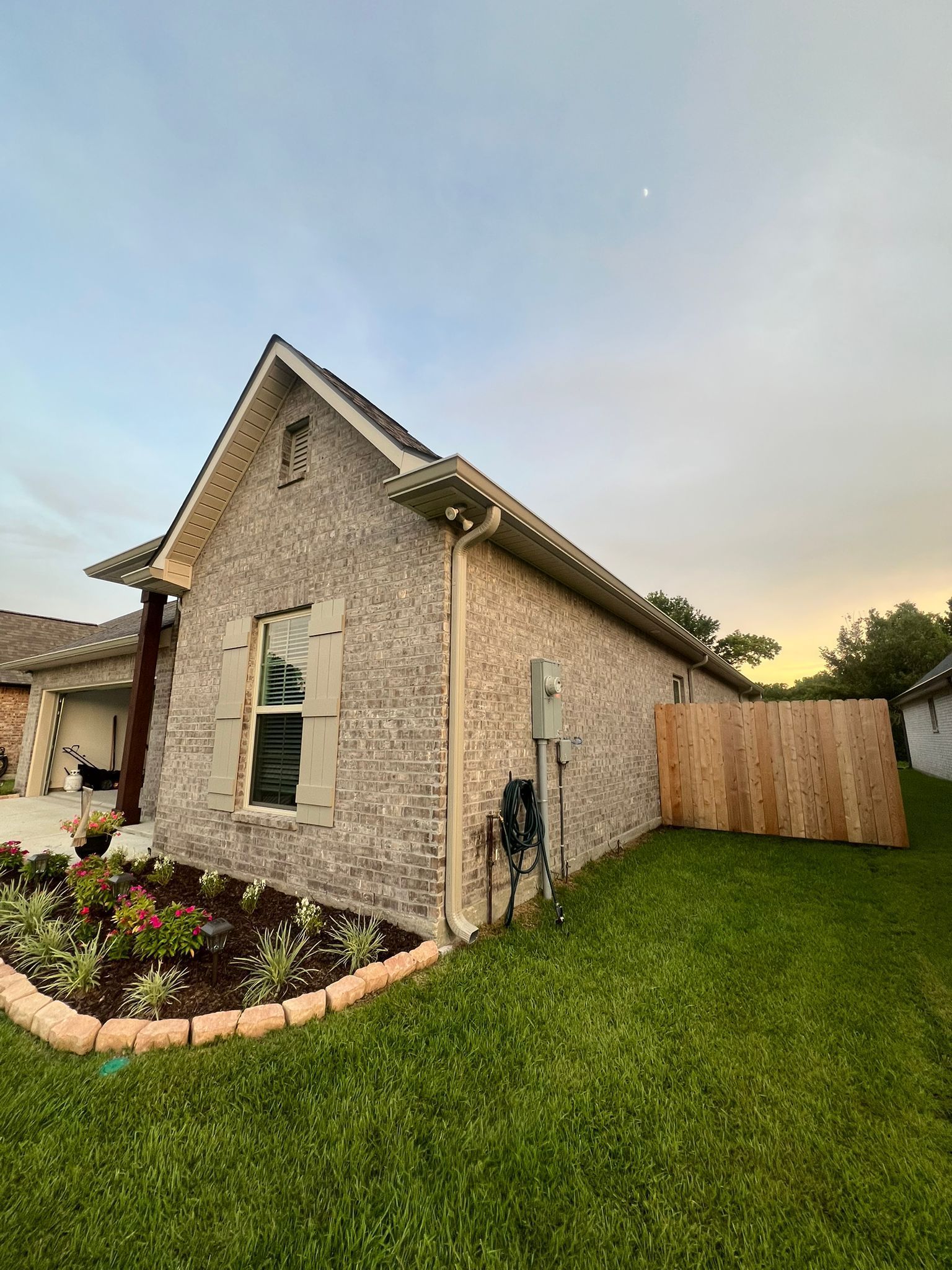 A single-story gray brick house with a gabled roof and a wooden fence on the side, set against a sunset sky.