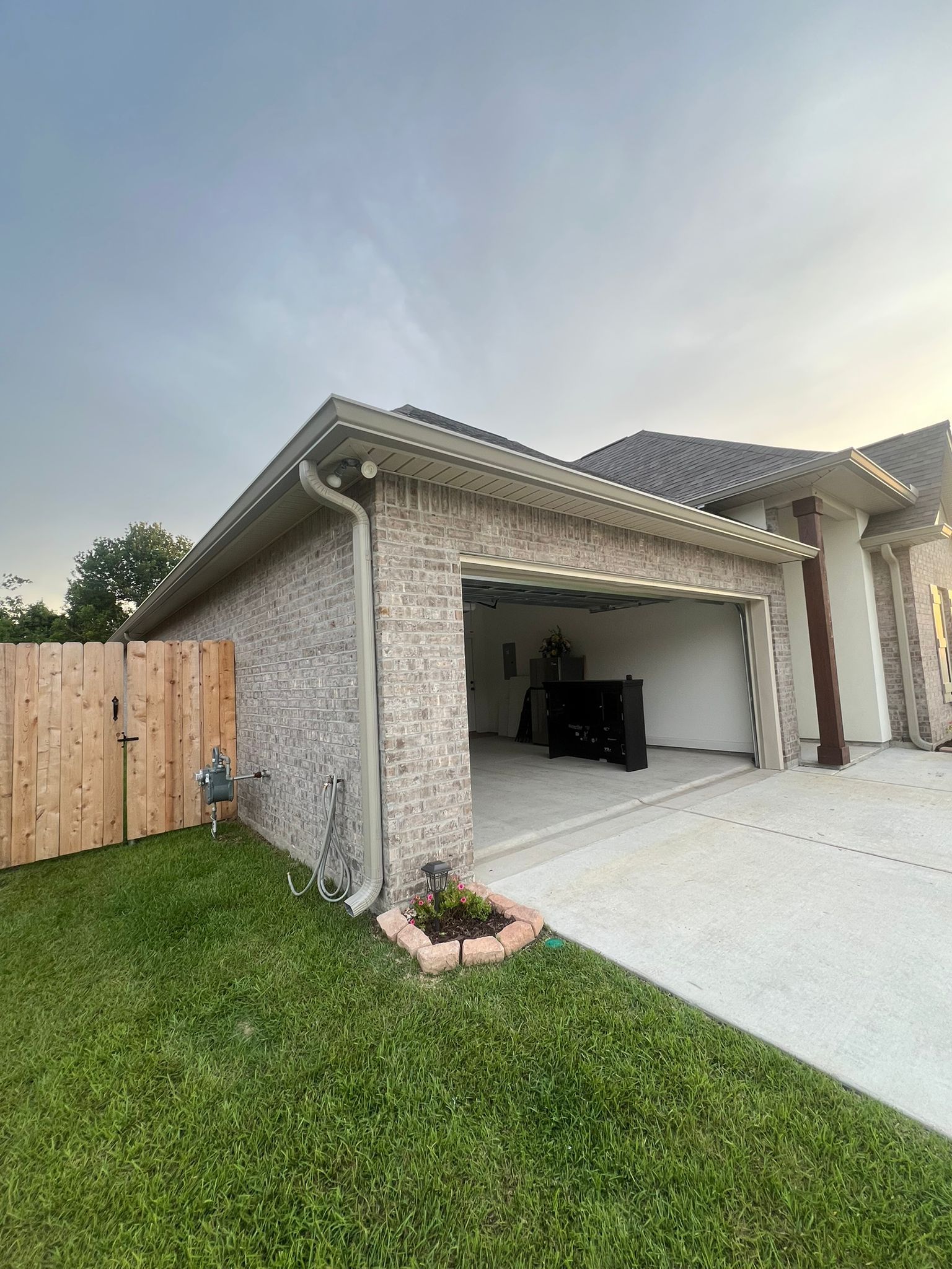 A side view of a stone-faced garage with an open door, next to a wooden fence and a grassy lawn under a pale sky.
