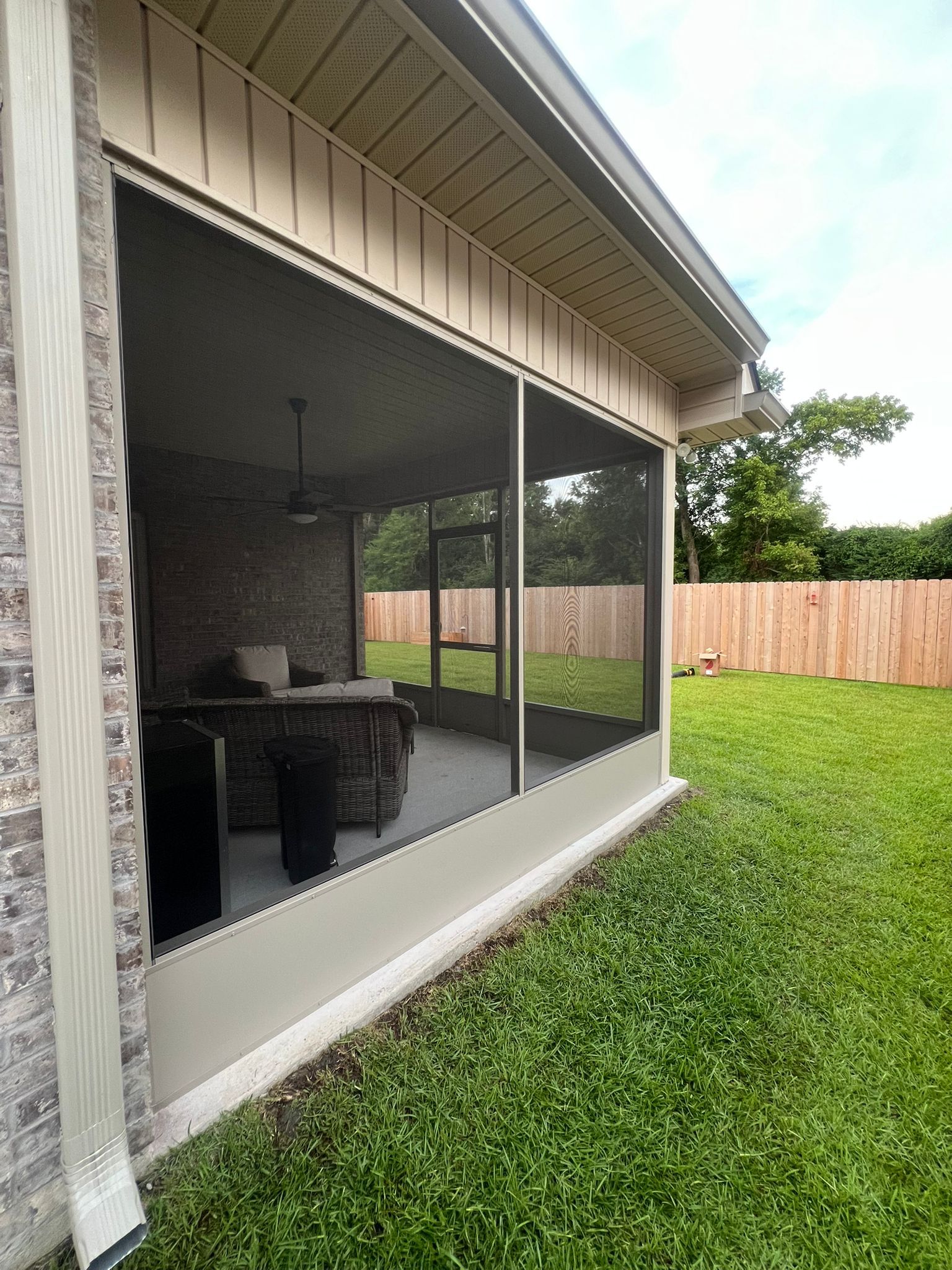 A screened-in patio with light-colored siding and a brick corner, viewed from a grassy backyard.