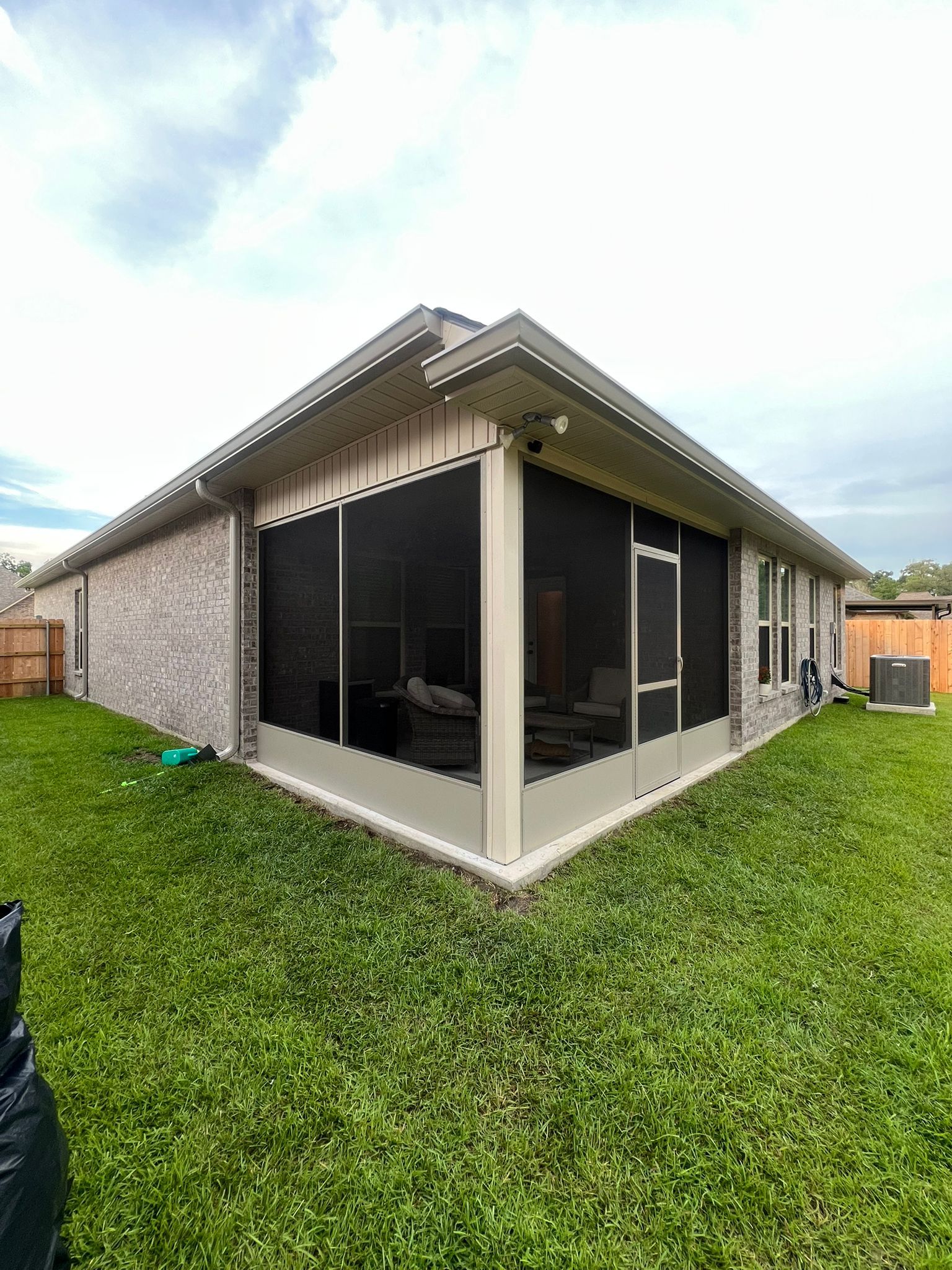 A corner view of a residential brick house featuring a tan-framed, screened-in porch, set in a green grassy backyard.