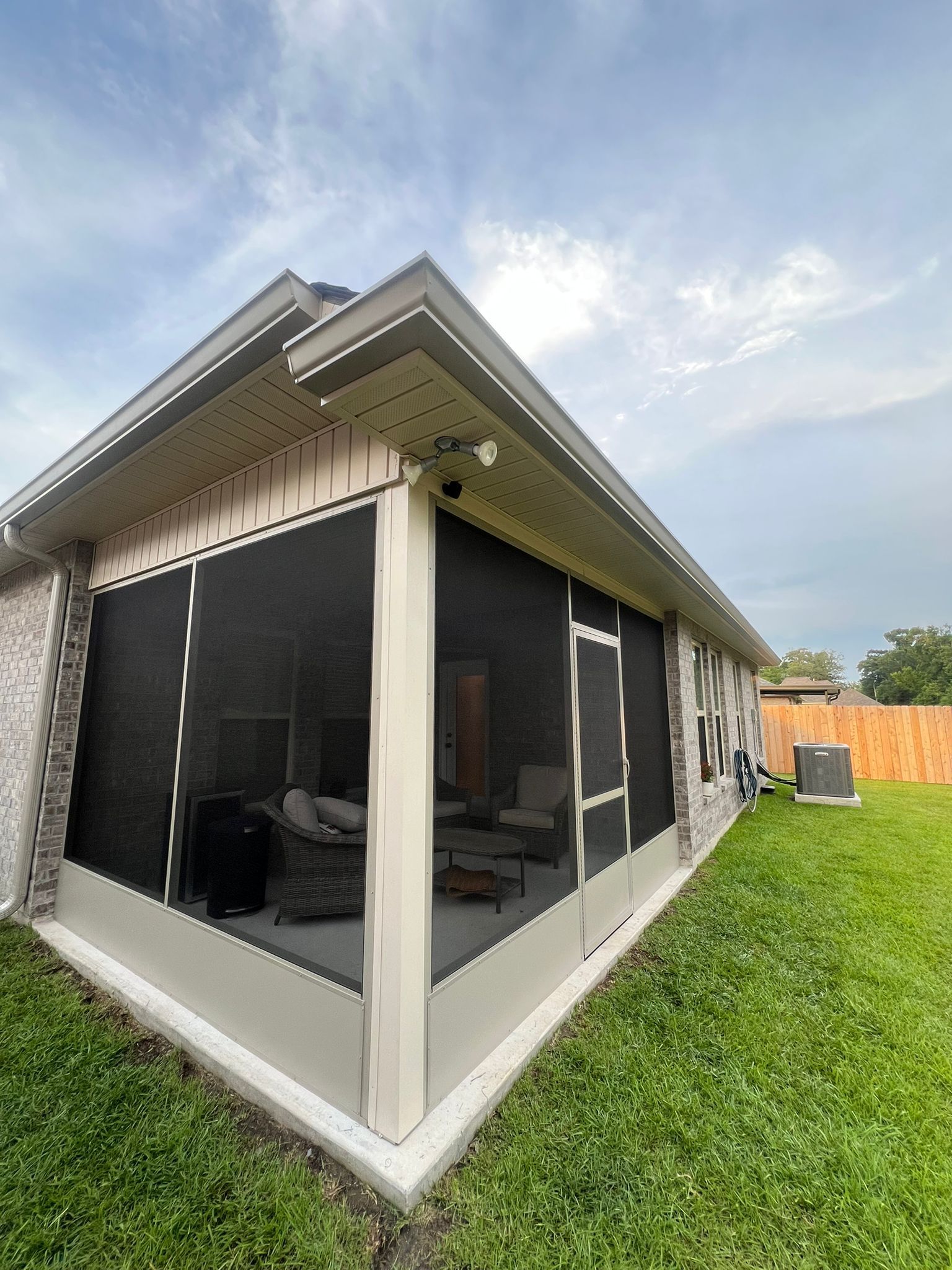 A tan screened-in patio with a gabled roof and white structural posts, viewed from a grassy backyard.