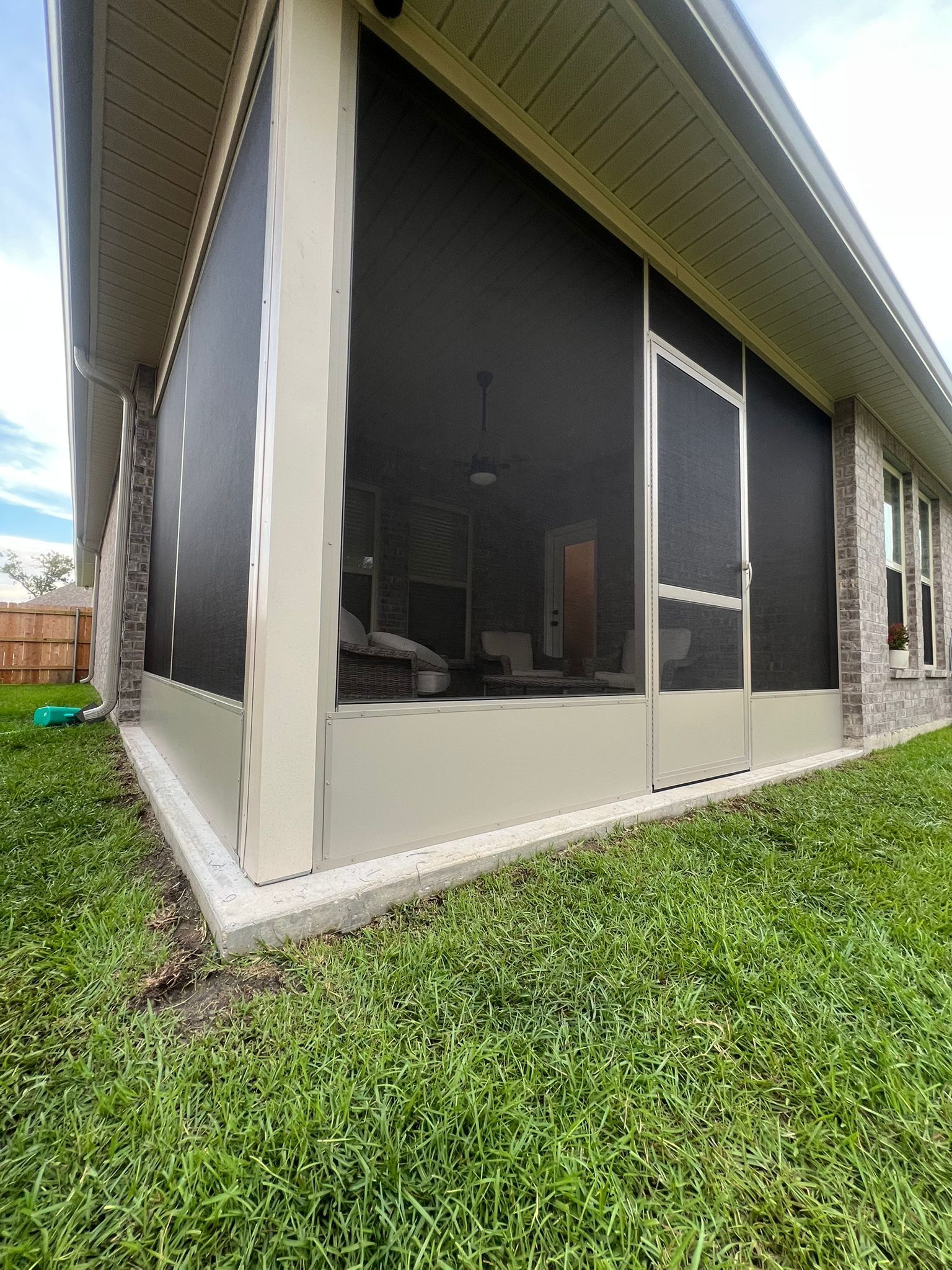 A beige, screened-in patio enclosure with a door, attached to a brick house with grass in the foreground.