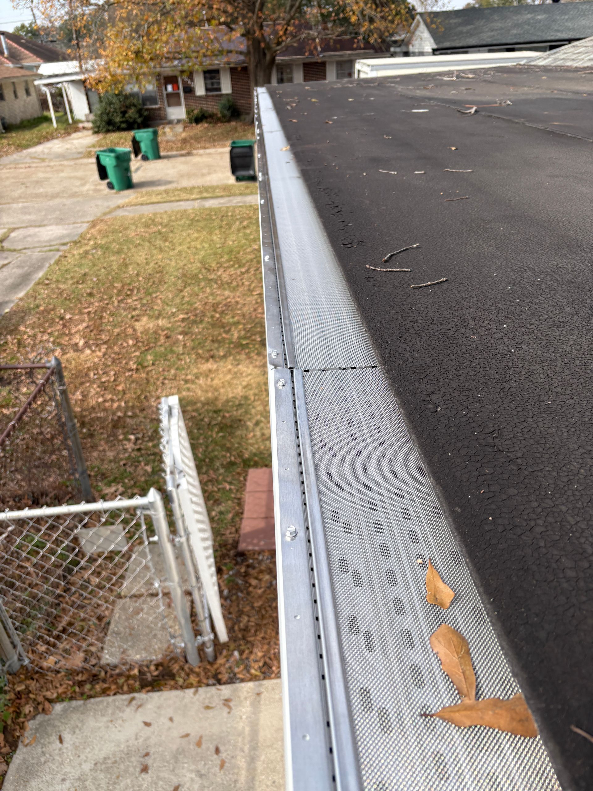 A metal gutter guard installed along the edge of a flat, dark-roofed building overlooking a residential yard.