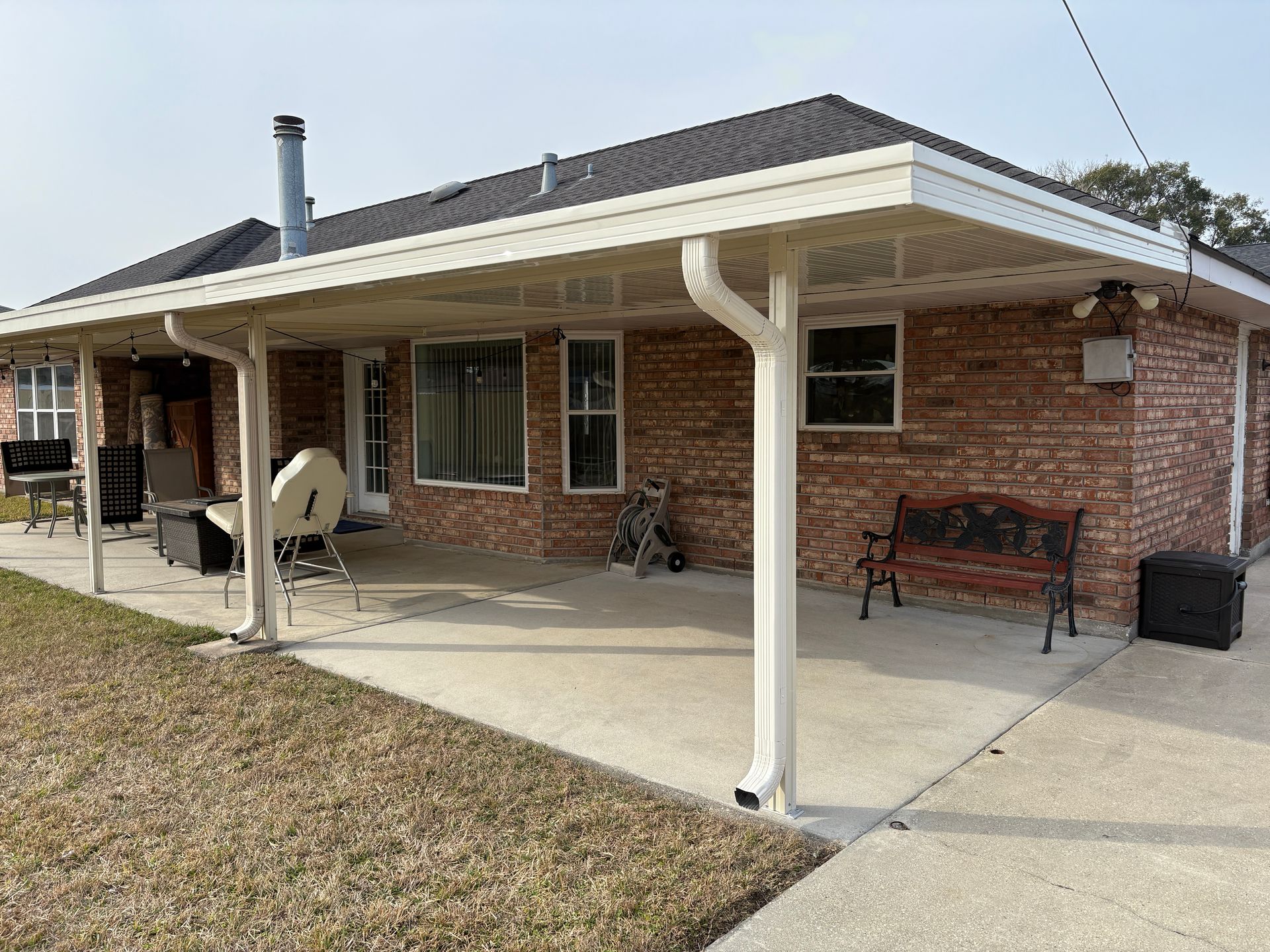 A brick house with a covered concrete patio, featuring white support posts, gutters, and outdoor seating.