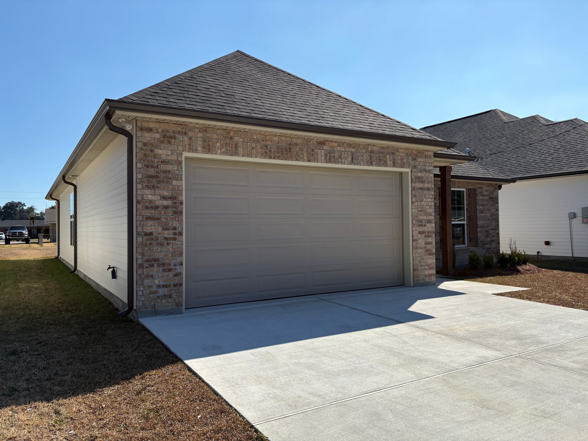 A tan, brick-front single-car garage with a closed door, a concrete driveway, and a shingled roof under a clear blue sky.