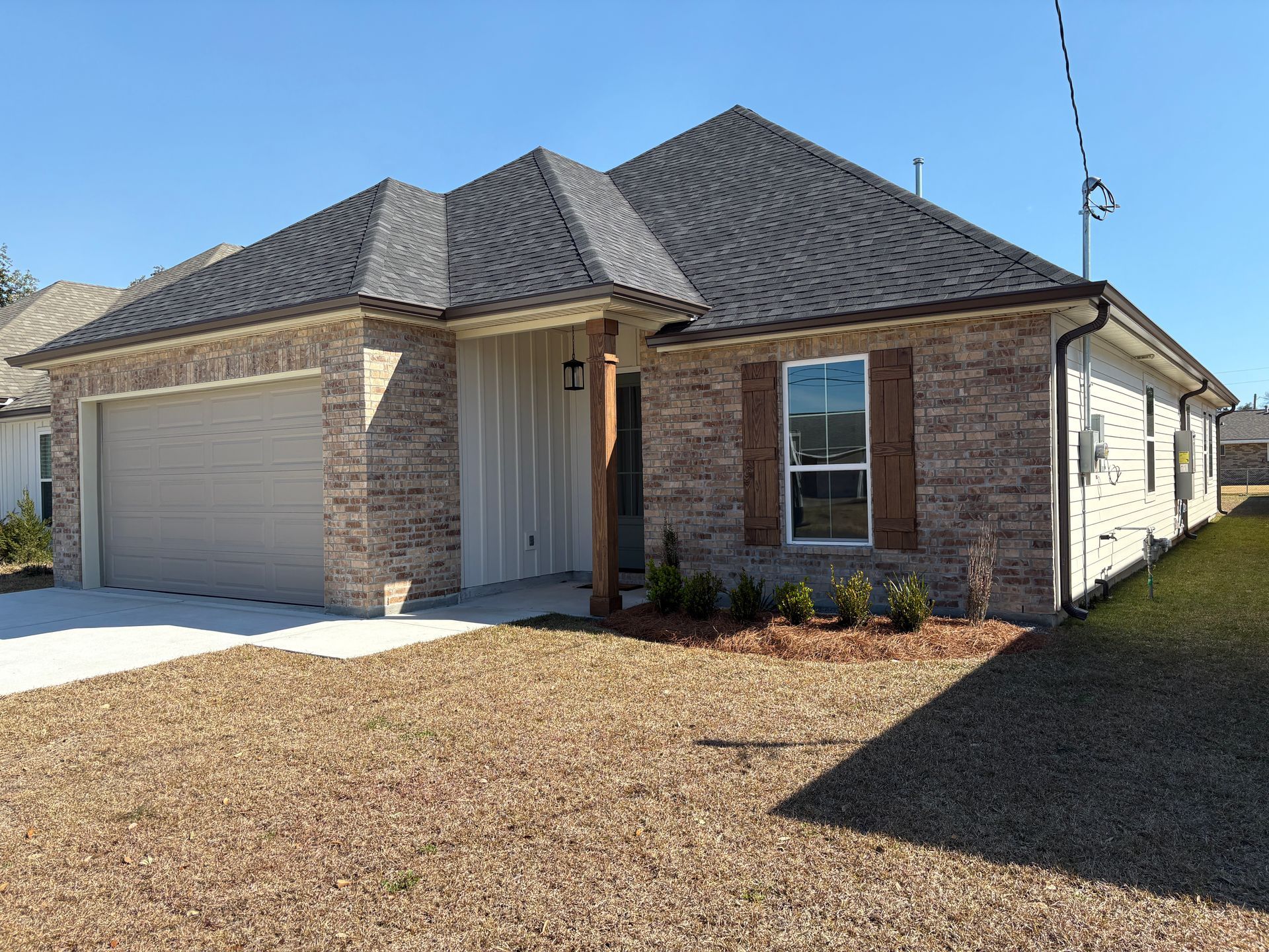 A single-story, brick-faced house with a two-car garage, grey shingled roof, and a wooden porch post under a blue sky.