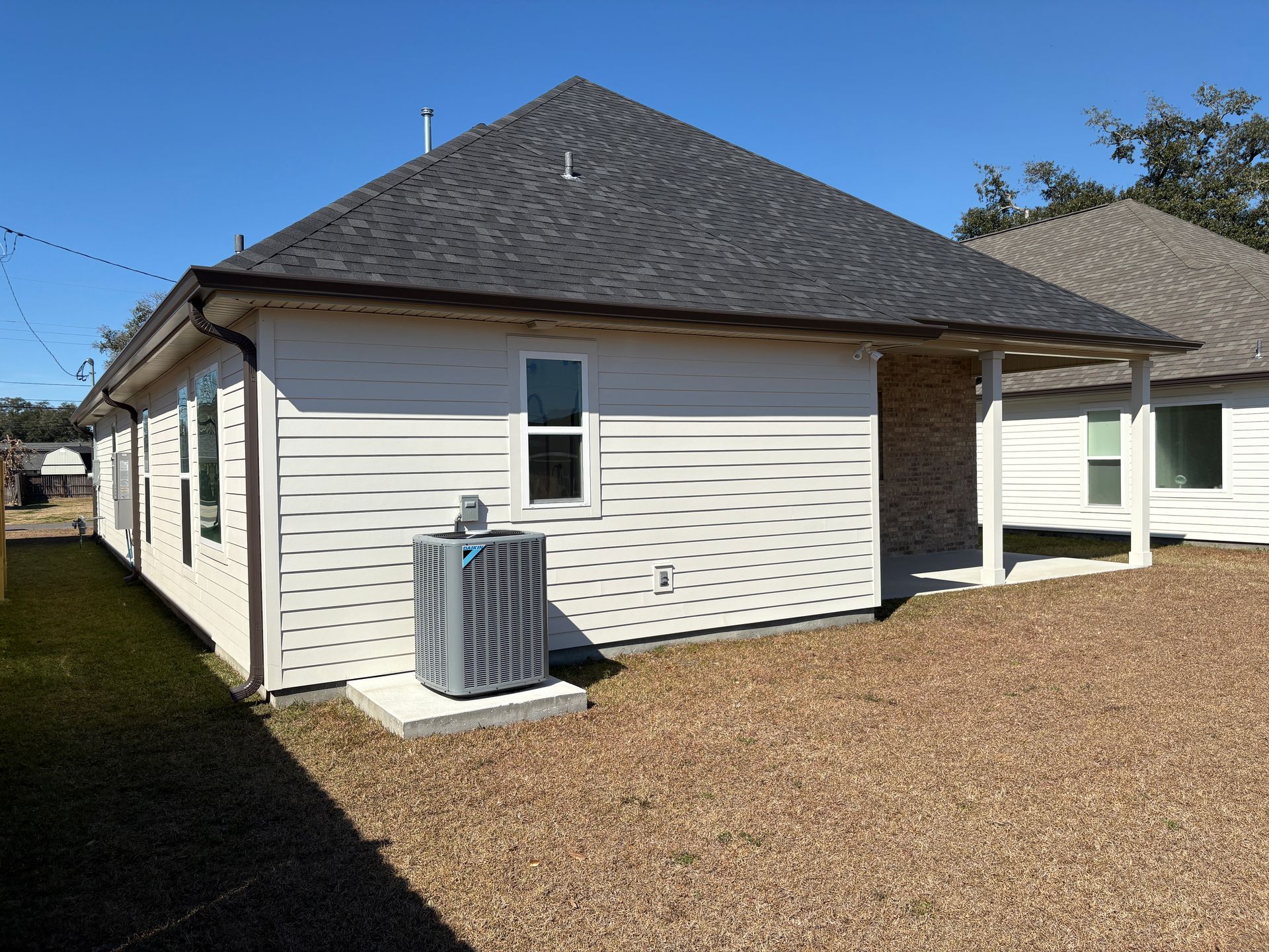 White house exterior with a gray shingle roof, outdoor AC unit on a concrete pad, and gravel yard under a blue sky.