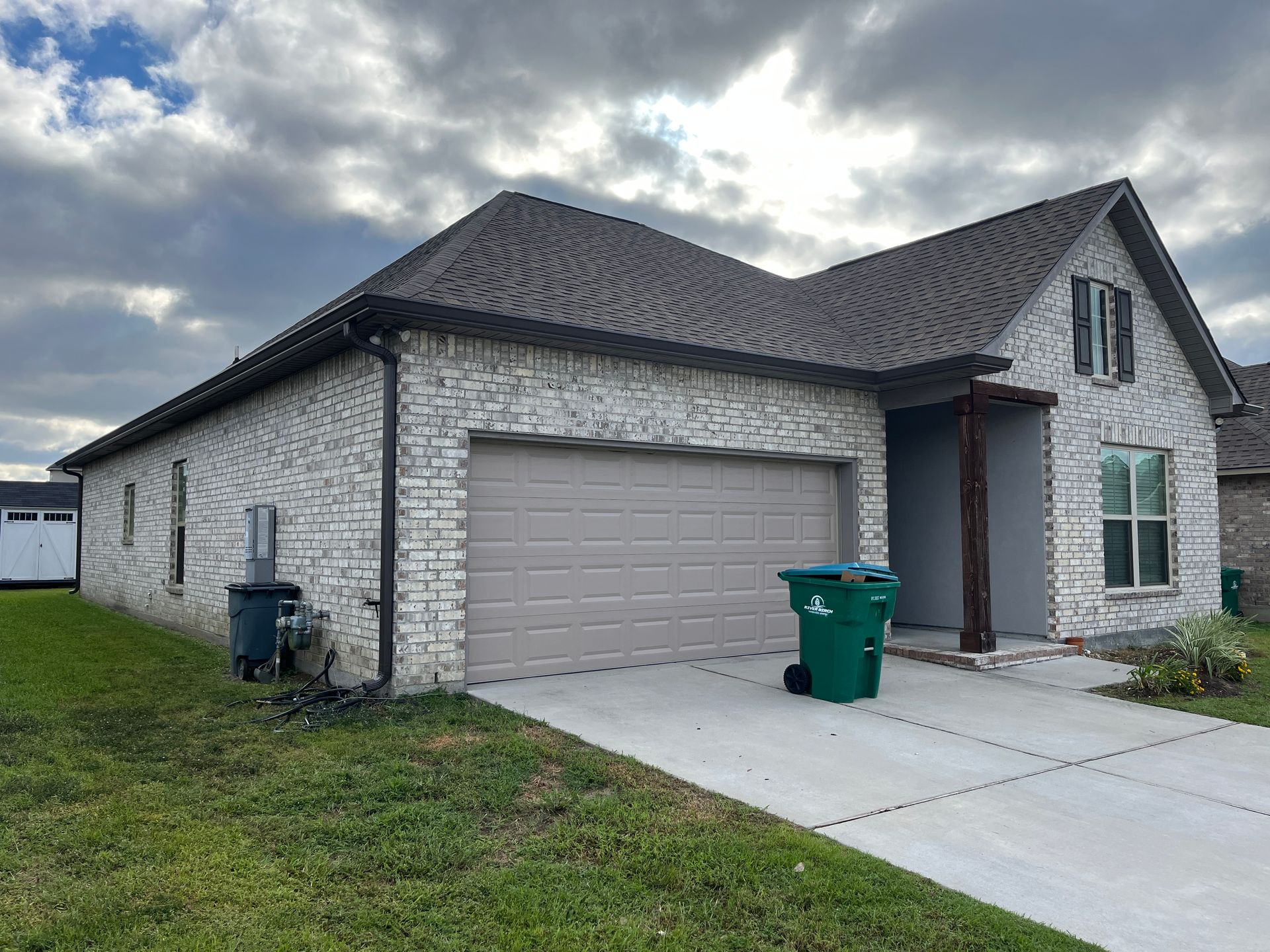 A light-colored brick, single-story suburban house with a two-car garage, gray roof, and a green trash bin on the driveway.