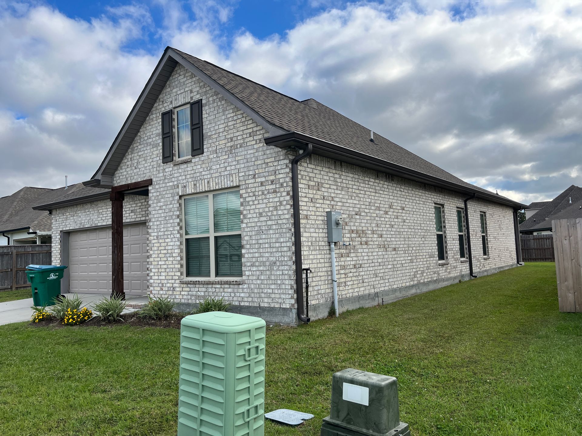 A side-angled view of a single-story white brick house with a dark roof, two-car garage, and green lawn under a cloudy sky.