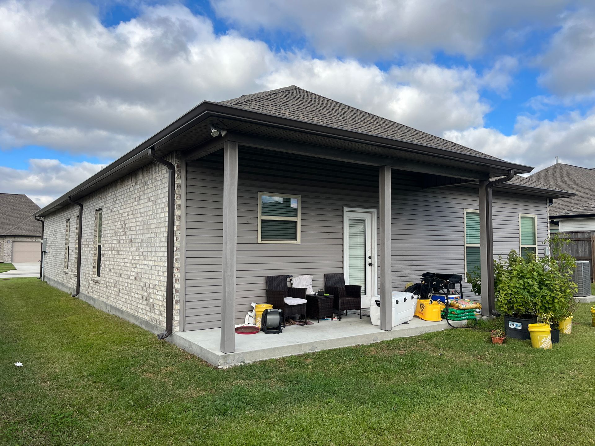 A backyard view of a single-story house with light stone siding and a covered gray patio with outdoor furniture.