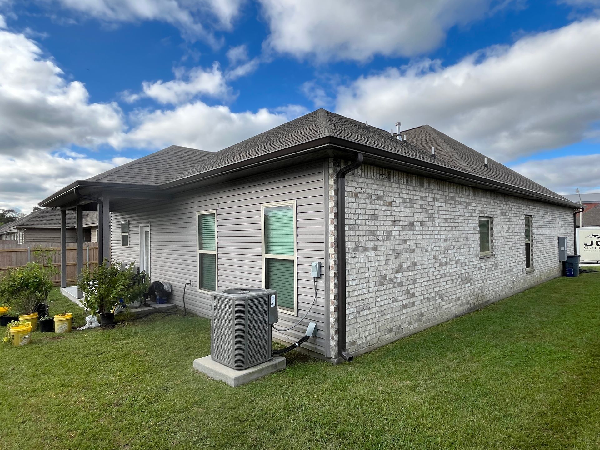 Back exterior view of a single-story brick house with a dark shingled roof, AC unit, and a small yard under a cloudy sky.