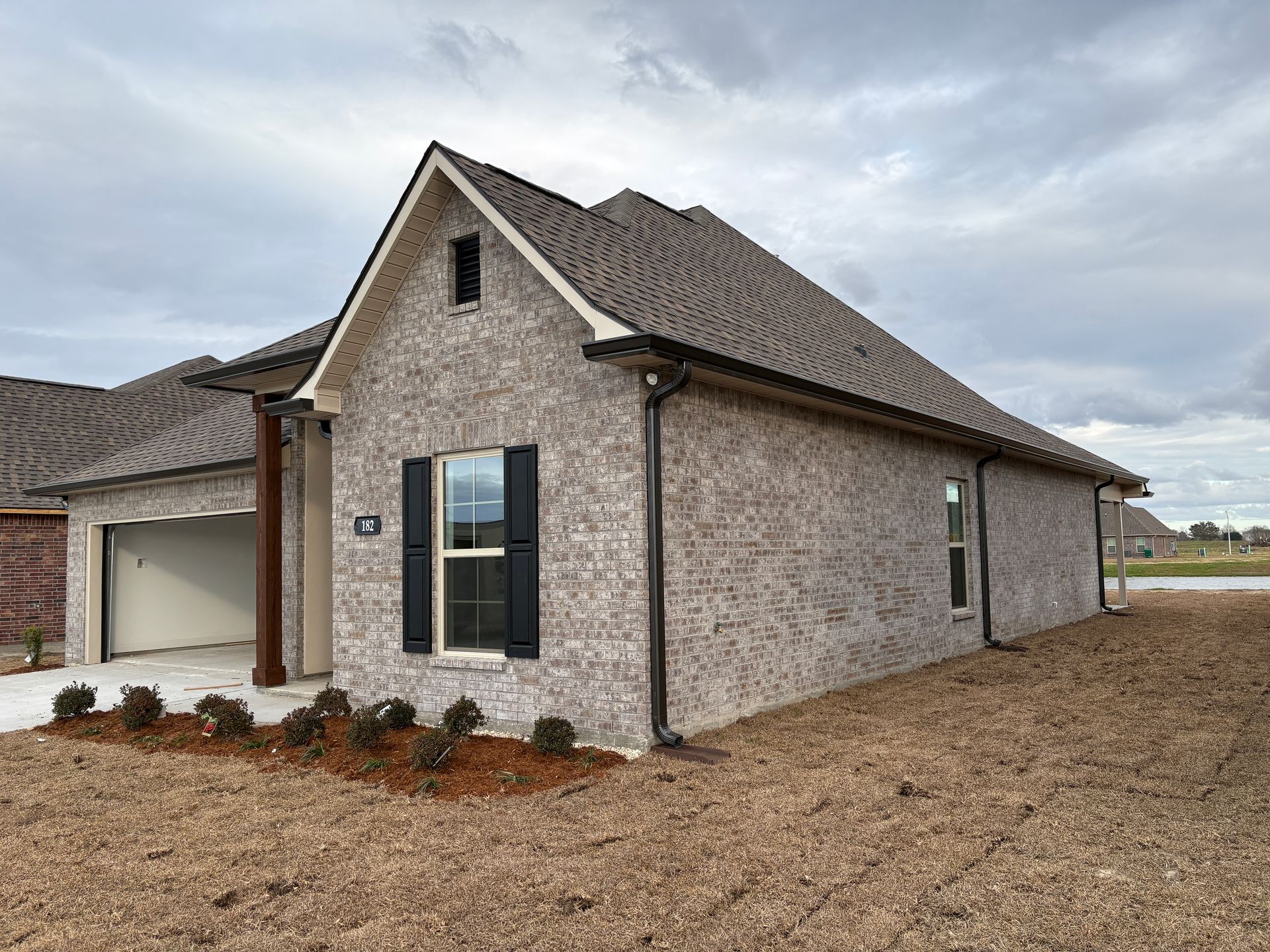 A single-story, grey brick house with a dark gabled roof, an open garage, black window shutters, and fresh landscaping.