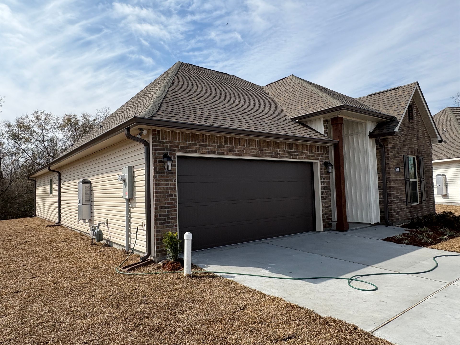 A tan-sided and brick house with a dark garage door, gray shingled roof, and a concrete driveway on a sunny day.