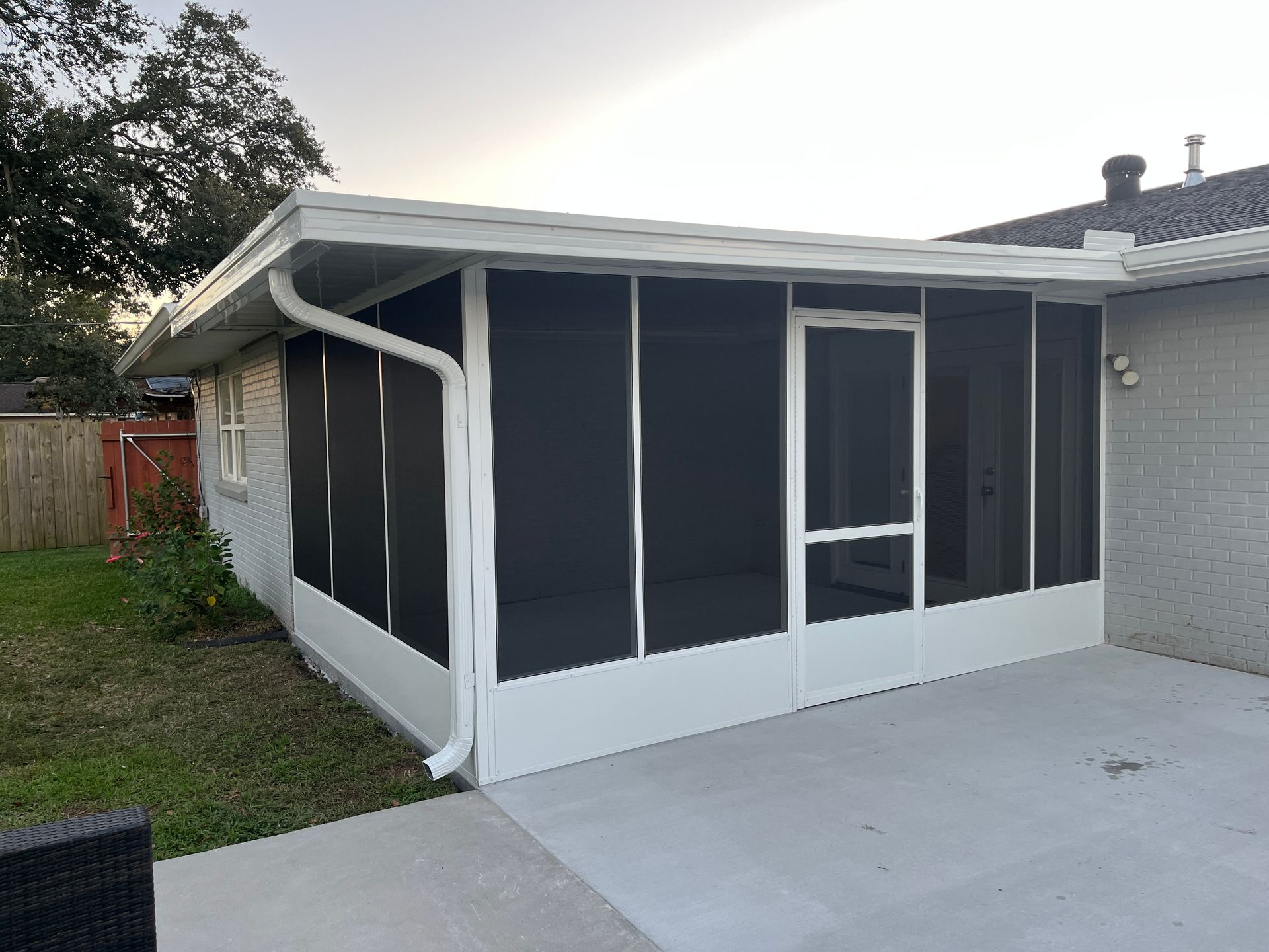 A white-framed screened-in porch attached to a light-colored brick house, featuring a white door and a concrete patio.