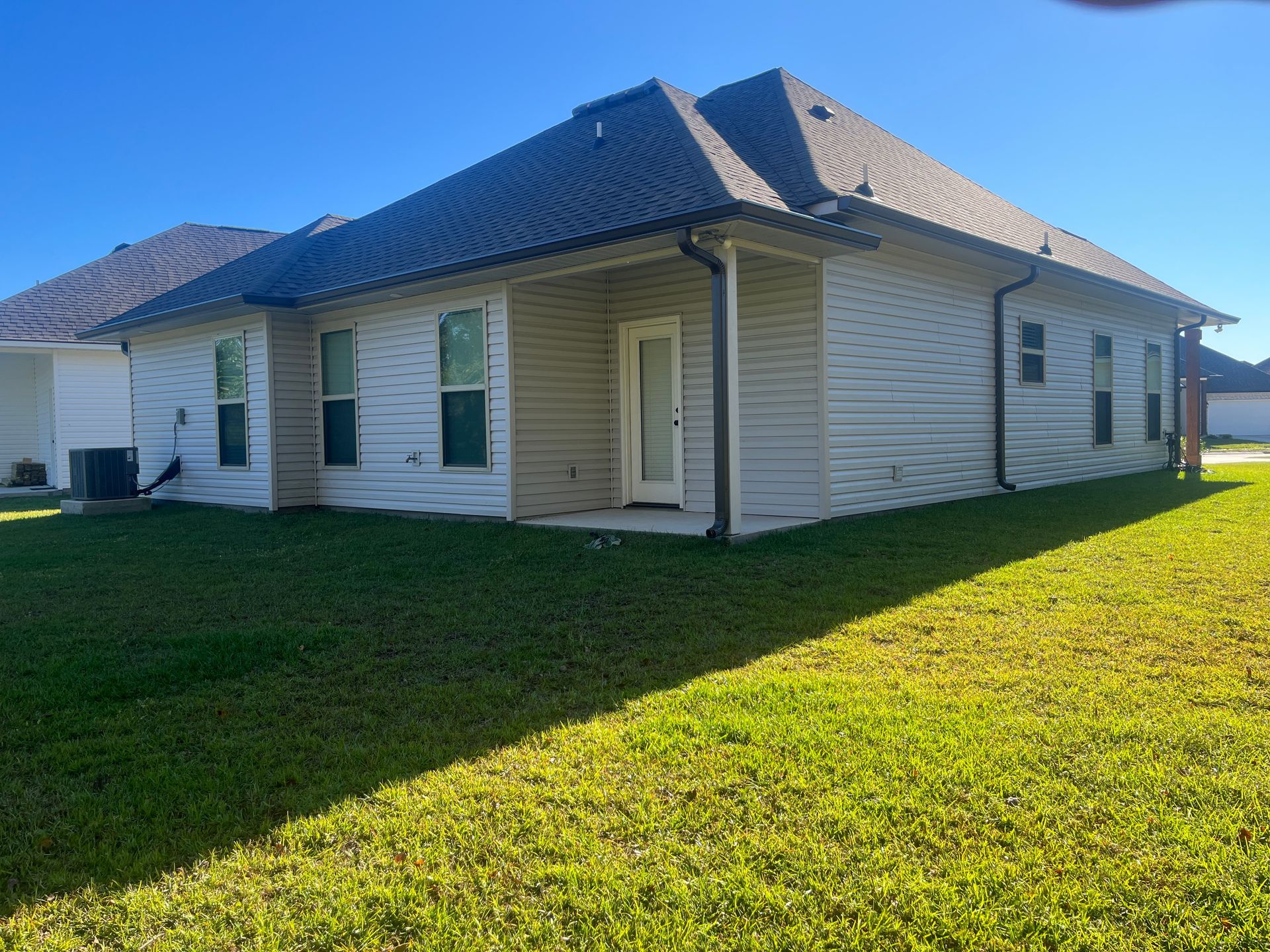 Back view of a single-story beige house with a dark roof and a lawn on a sunny day.