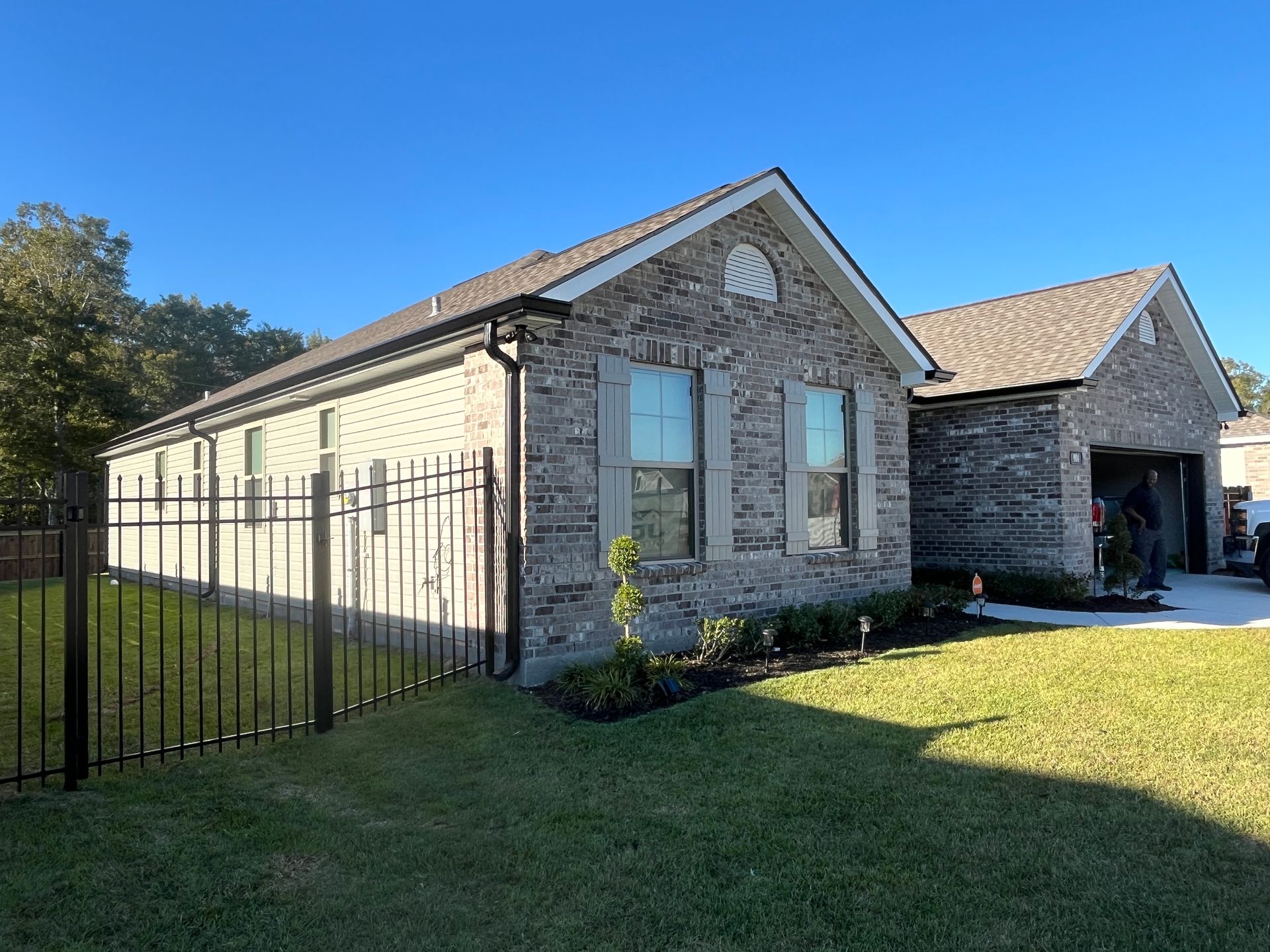 A single-story brick house with a two-car garage, a black metal fence, and a front lawn under a clear blue sky.