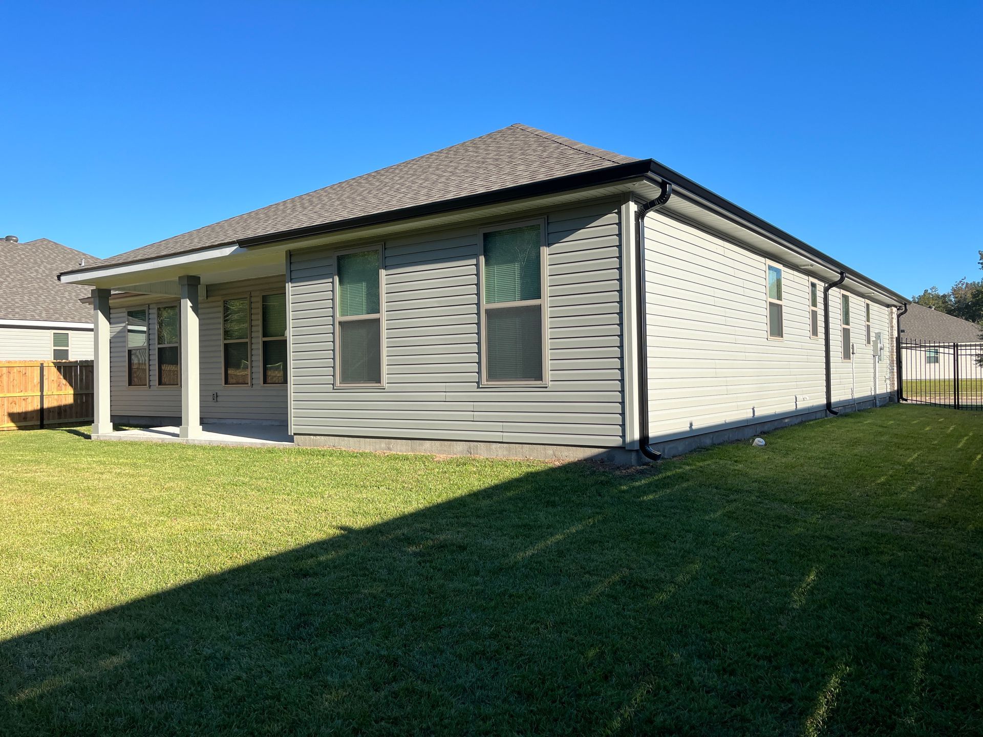 A light-colored house with horizontal siding and a covered back patio faces a grassy lawn under a clear blue sky.