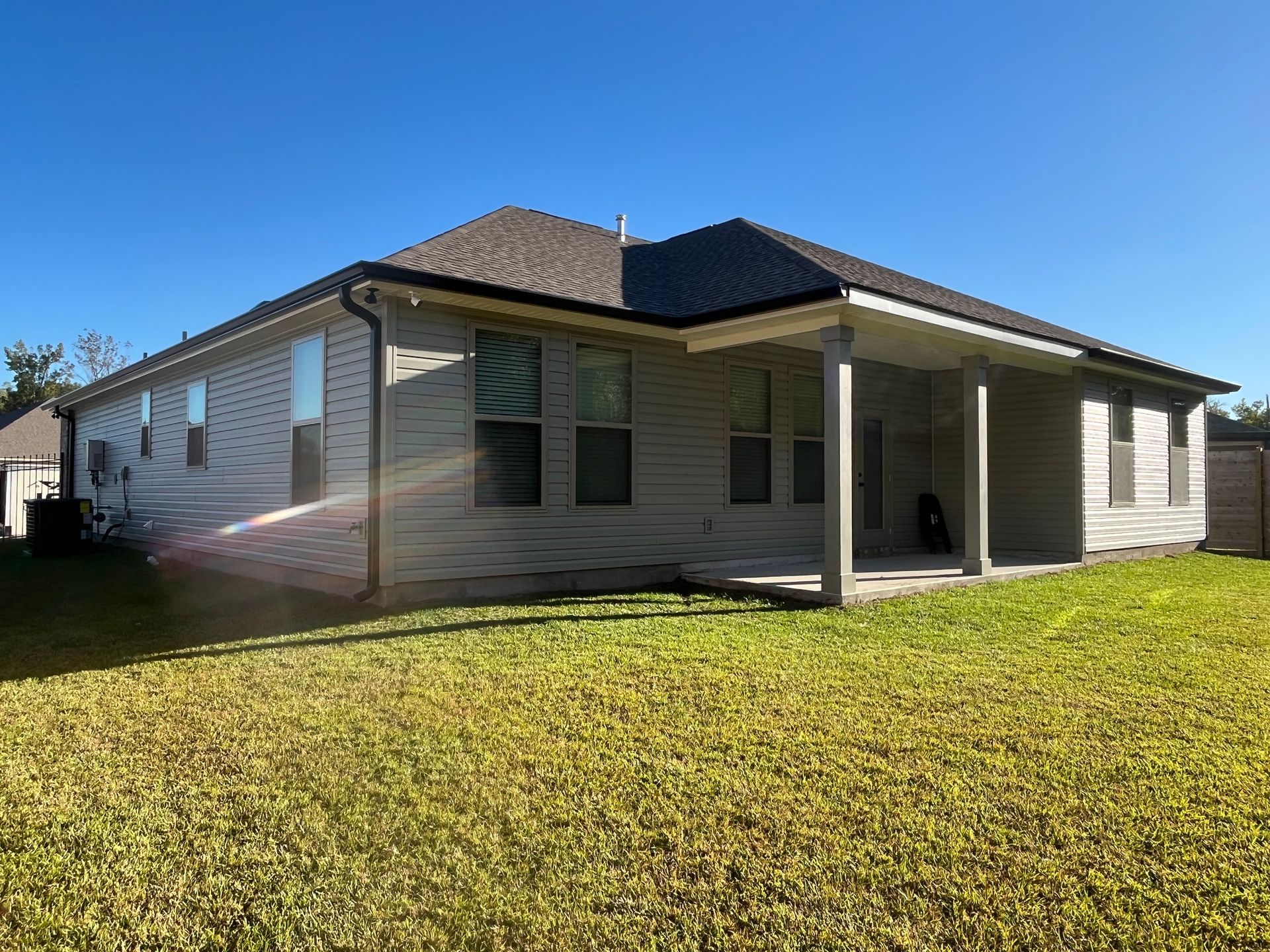 A one-story tan house with a covered back patio and shingled roof, surrounded by a large green lawn under a clear blue sky.