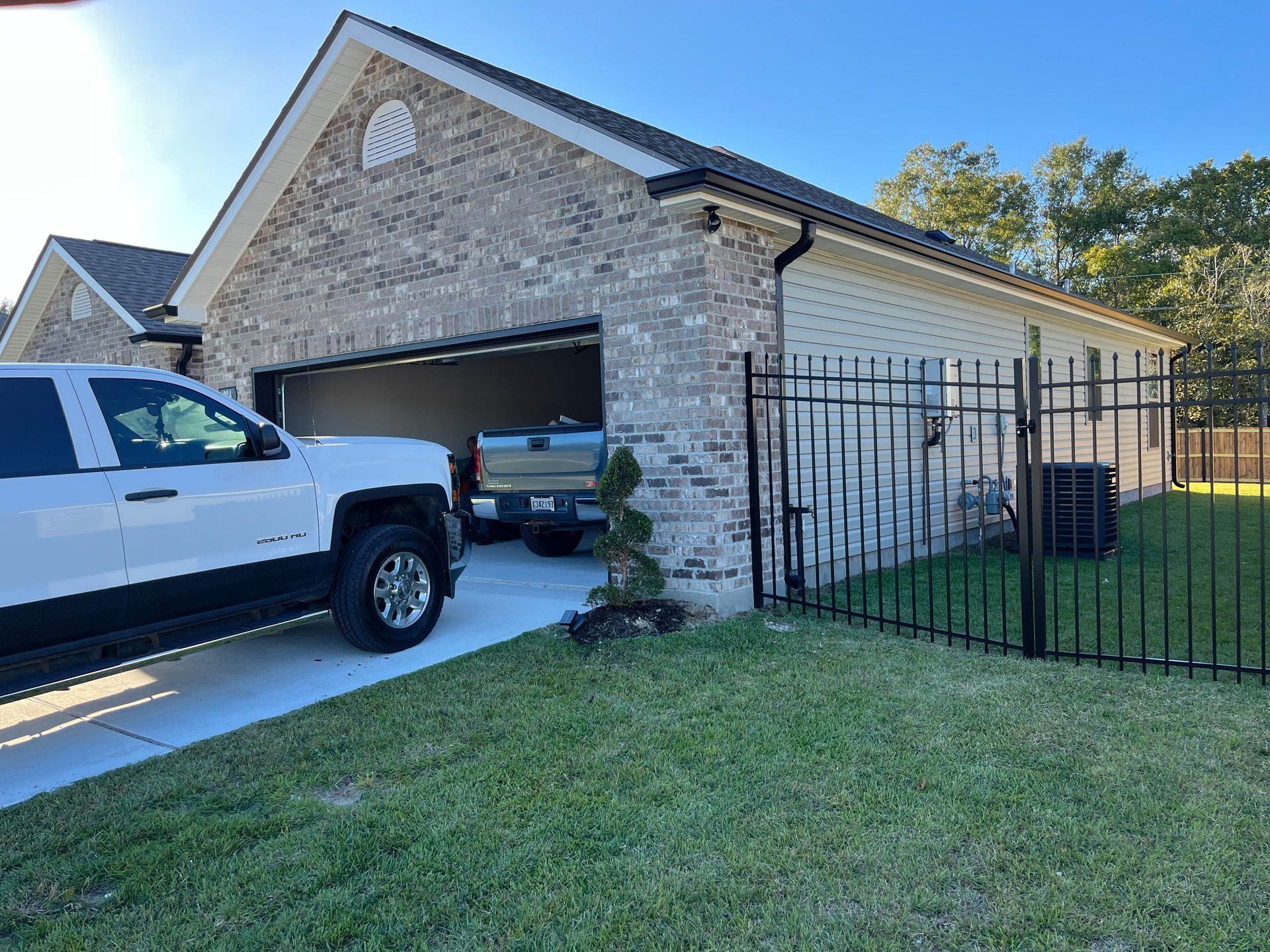 A white pickup truck parked in the driveway of a brick house with an open garage and a black metal fence on a sunny day.