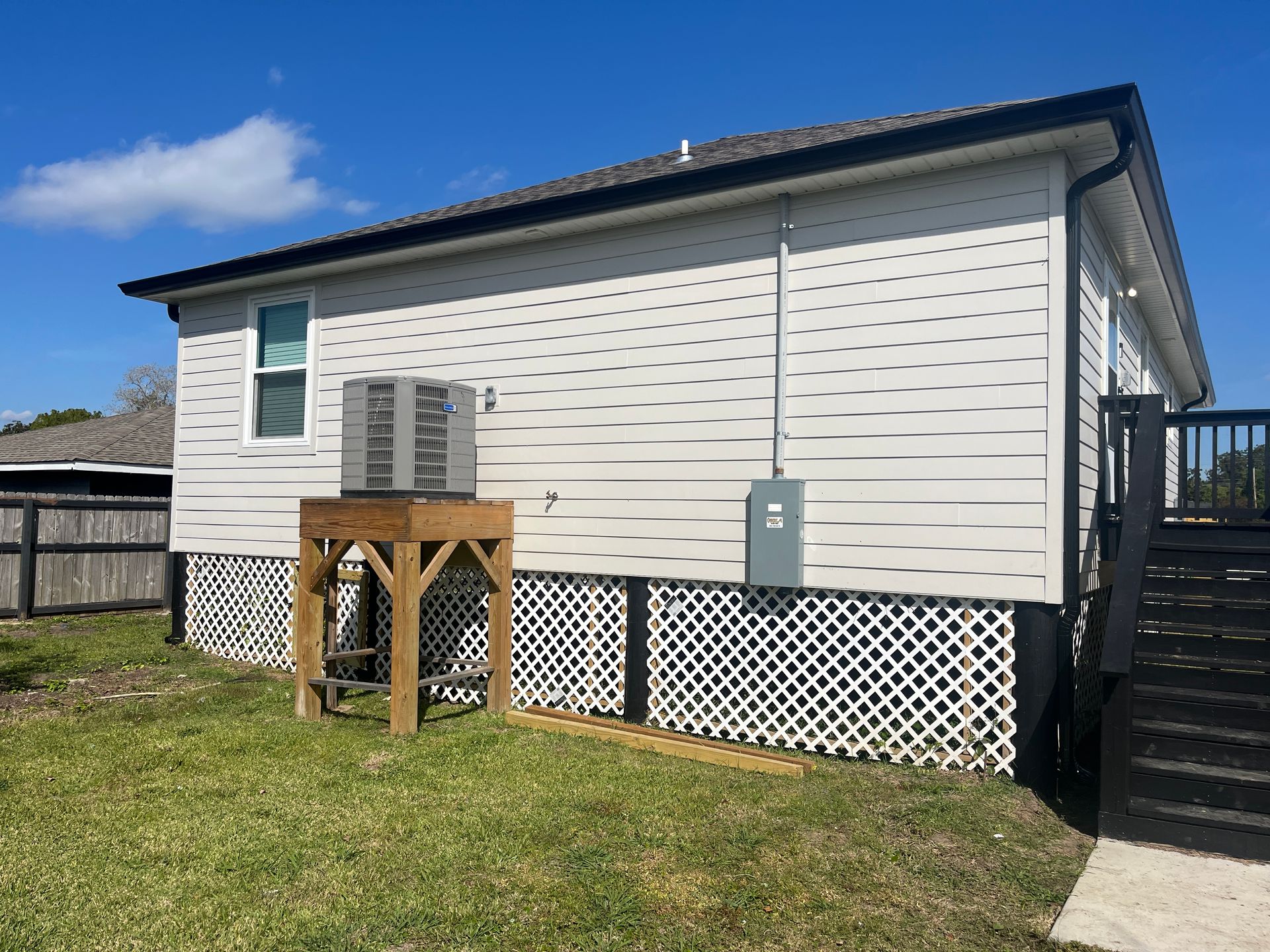 An exterior view of a beige siding house with an HVAC unit on a wooden stand and a black staircase on a sunny day.
