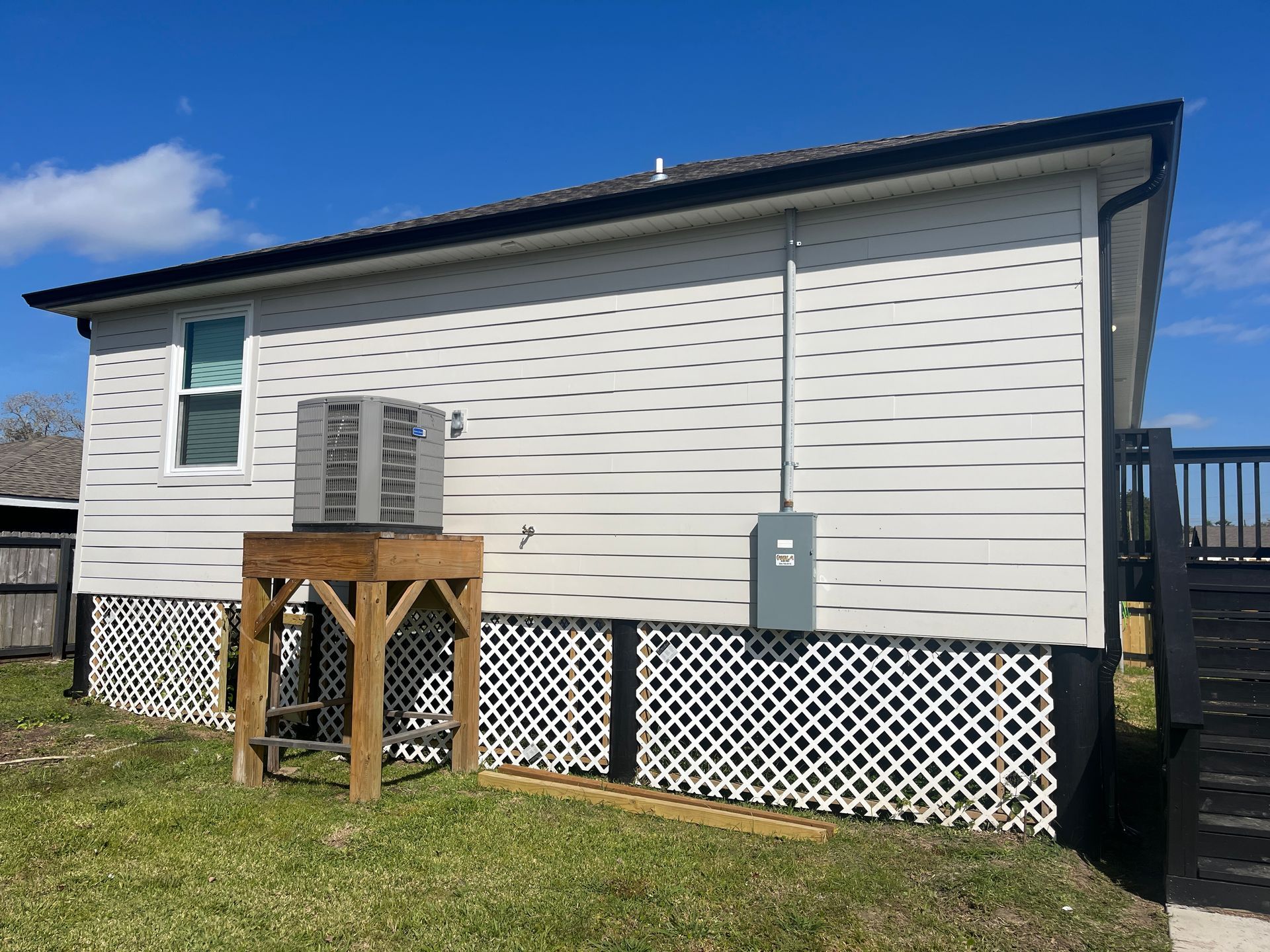 A beige house exterior with white lattice skirting, an AC unit on a wooden stand, and an electrical box on a sunny day.
