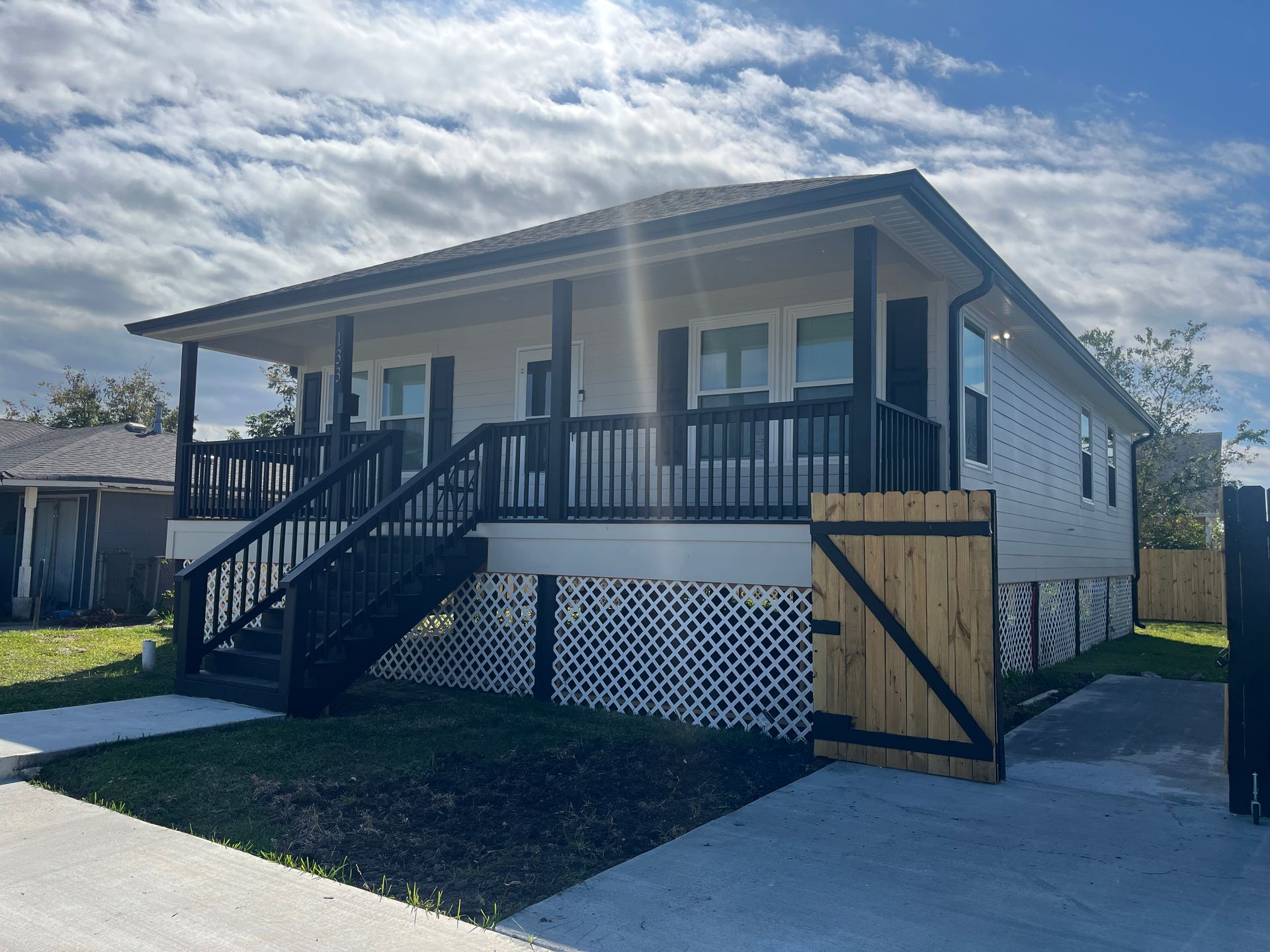 A single-story, light-colored house with a dark railing porch, raised foundation with lattice, and a wooden side gate.