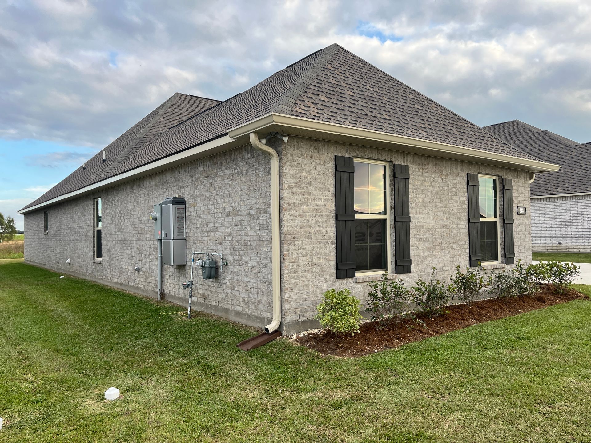 A side view of a light grey brick single-story house with black shutters, a dark roof, and a small patch of mulch.