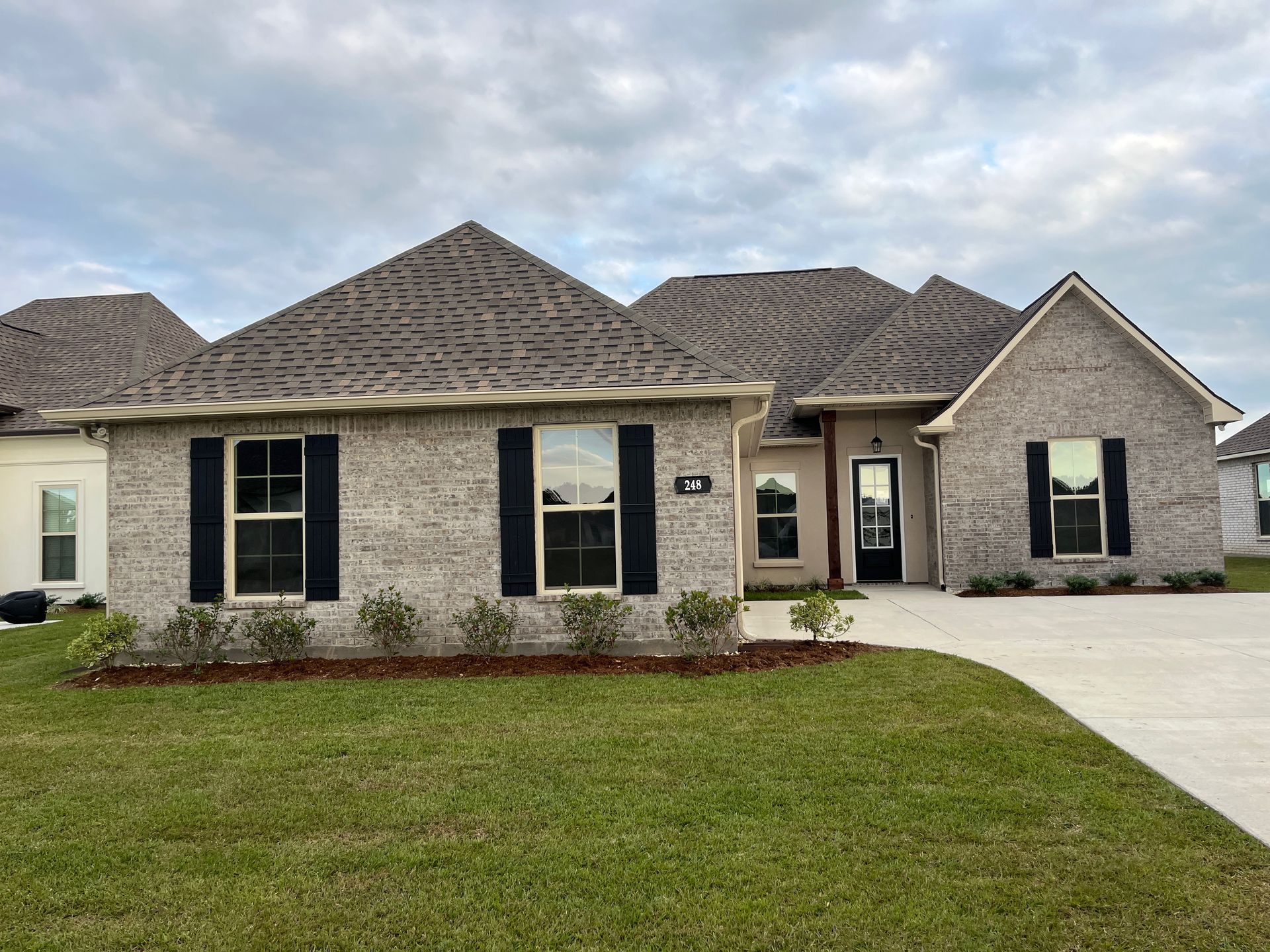 A single-story gray brick house with dark shutters, a front driveway, and a lawn under a cloudy sky.