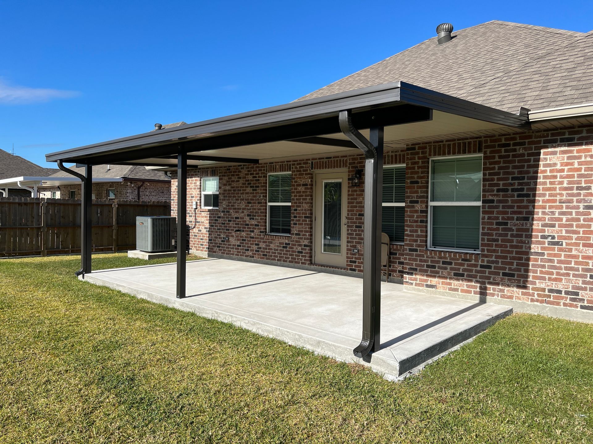 A brown brick house with a covered concrete patio featuring a dark metal roof supported by two black pillars.