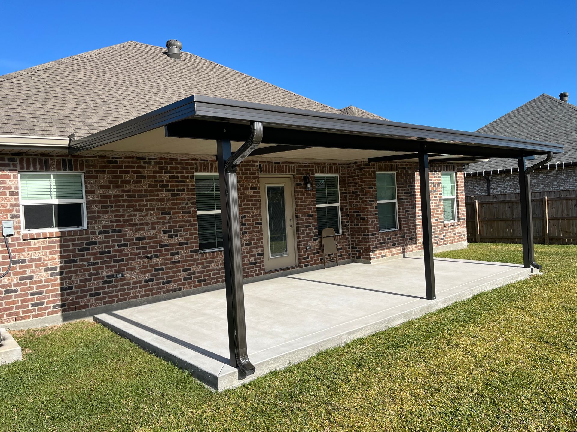 A brick house with a dark metal patio cover extending over a concrete patio in a grassy backyard under a clear blue sky.