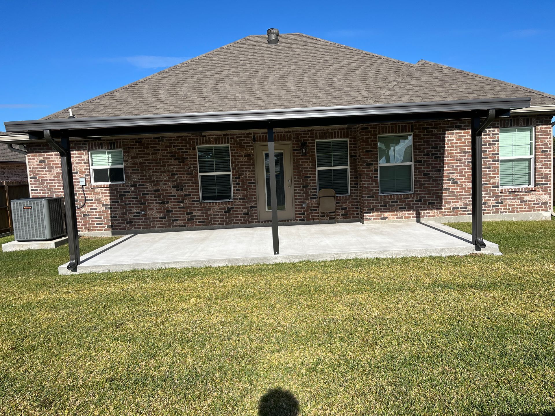 A brick house exterior features a concrete patio covered by a dark metal pergola under a clear blue sky.