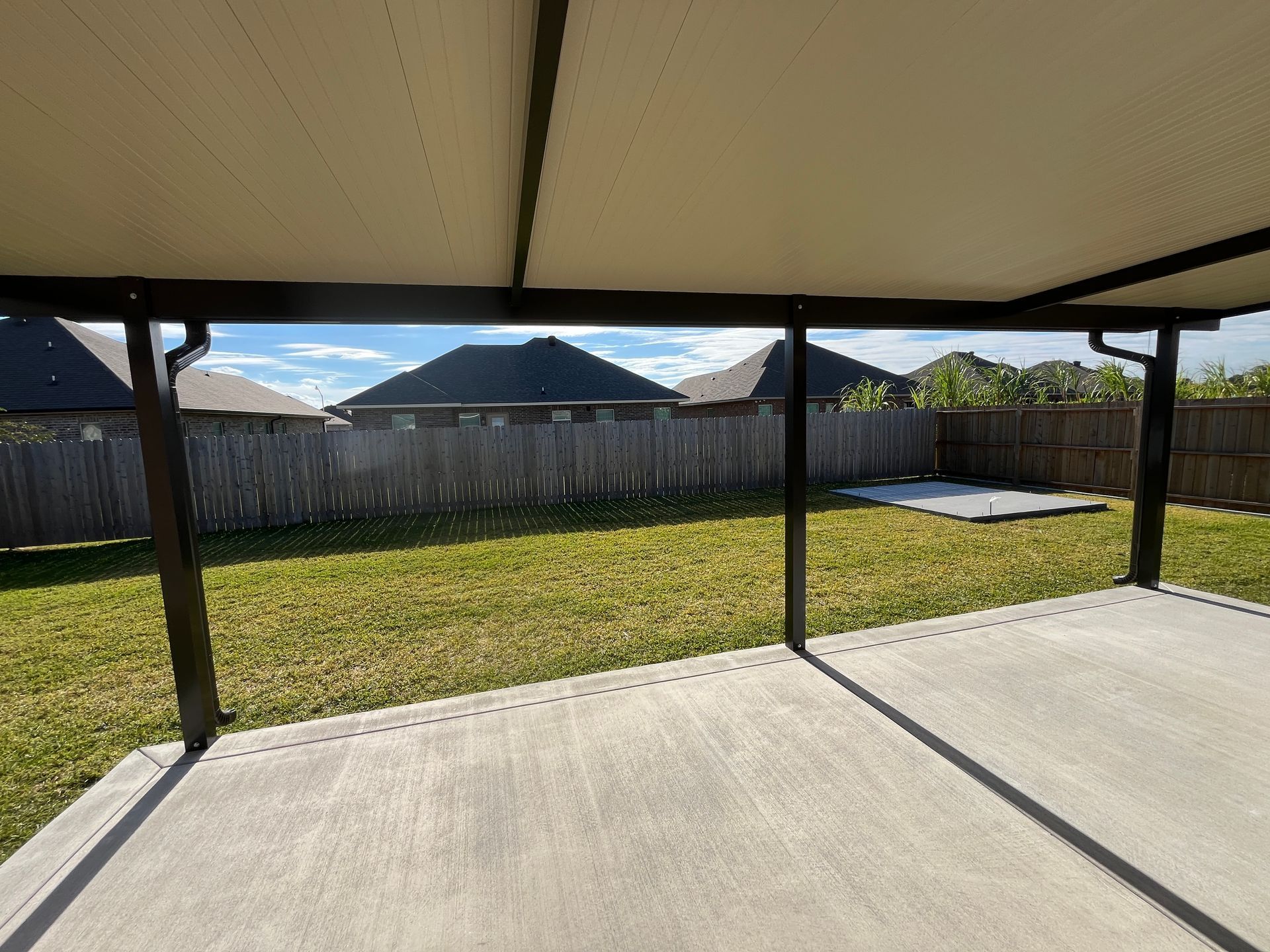 A view from a covered concrete patio looking out over a green backyard with a wooden fence and houses in the distance.