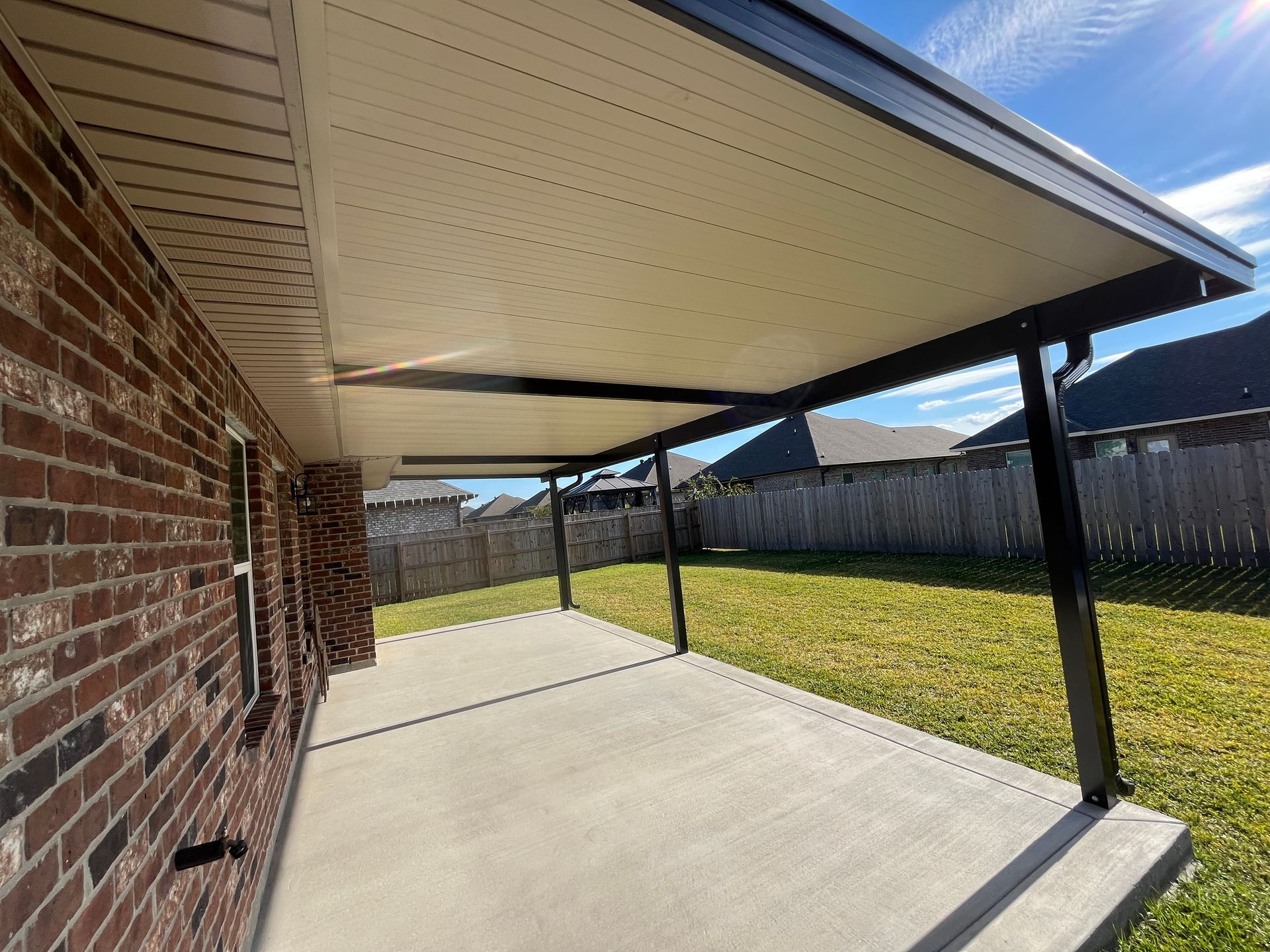 A covered backyard patio with a concrete floor, dark support posts, and a brick house wall.