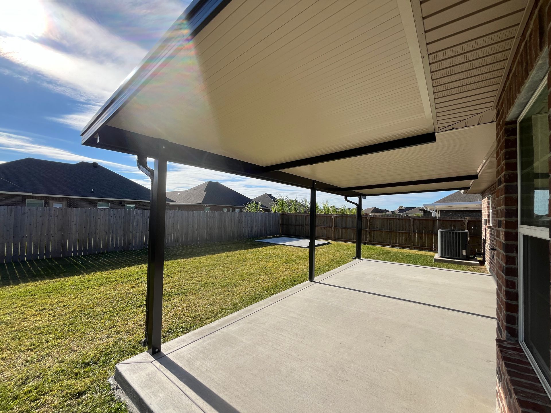 A covered patio with a concrete floor, dark support beams, and a light-colored ceiling looking out into a fenced backyard.