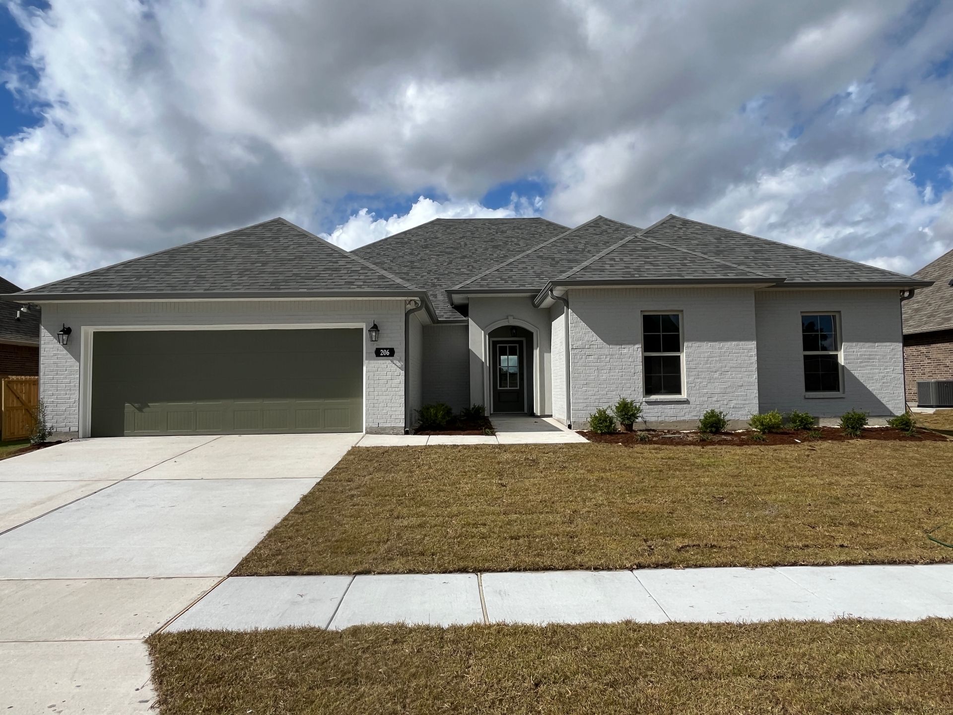 A single-story, light gray house with a two-car garage, a shingled roof, and a front lawn under a cloudy blue sky.