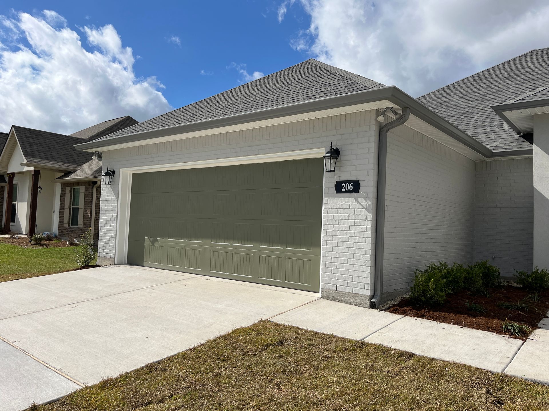 A view of a white brick suburban house with a large, olive green garage door and a gray shingled roof under a blue sky.