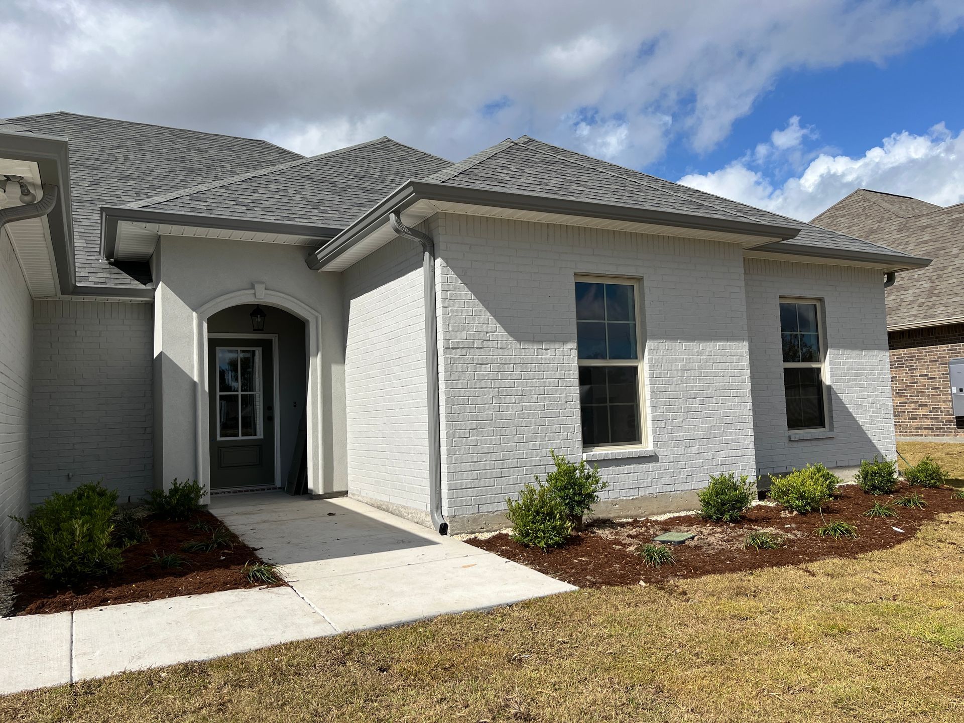 A single-story, light gray brick house with a front porch, concrete walkway, and landscaped beds under a cloudy blue sky.