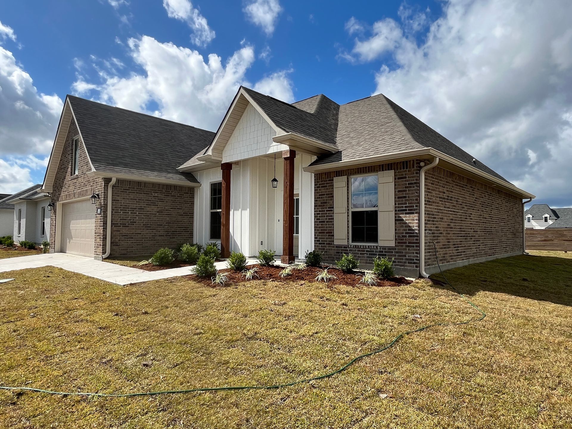 A single-story brick house with a brown shingled roof, white trim, and a small front garden under a partly cloudy blue sky.