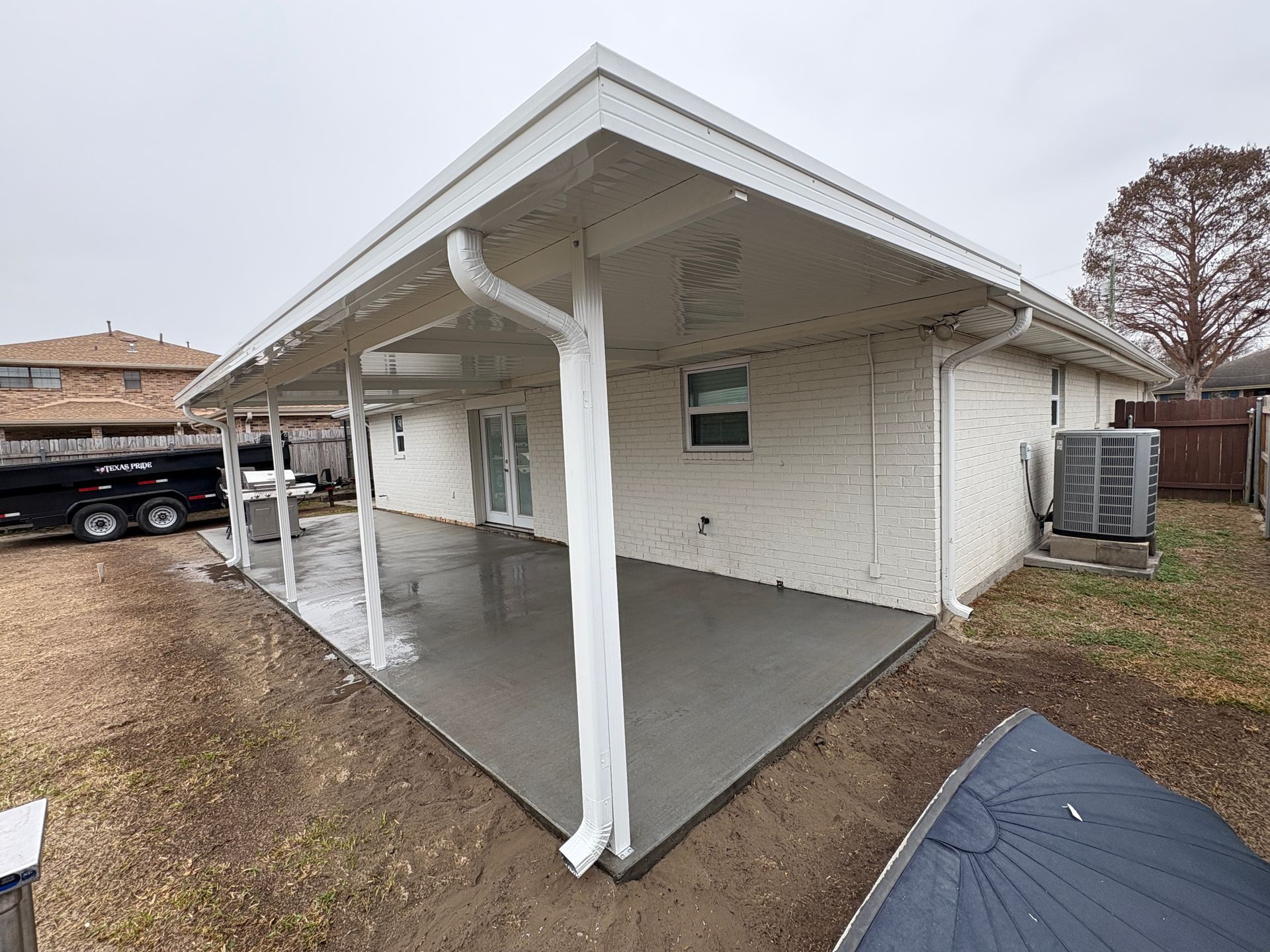 Newly poured concrete patio under a covered white porch attached to the back of a brick house.