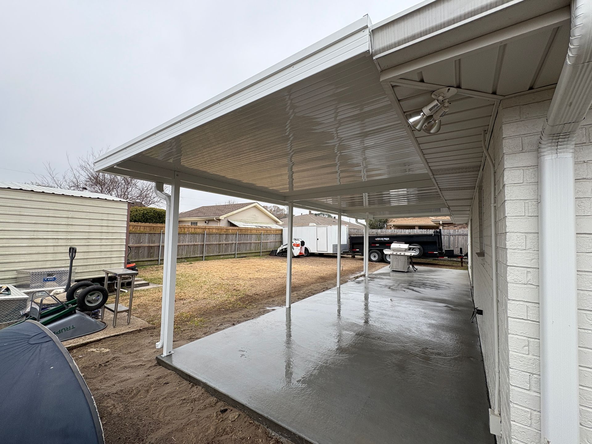 A newly poured concrete patio covered by a white metal roof attached to a white brick house in a backyard.