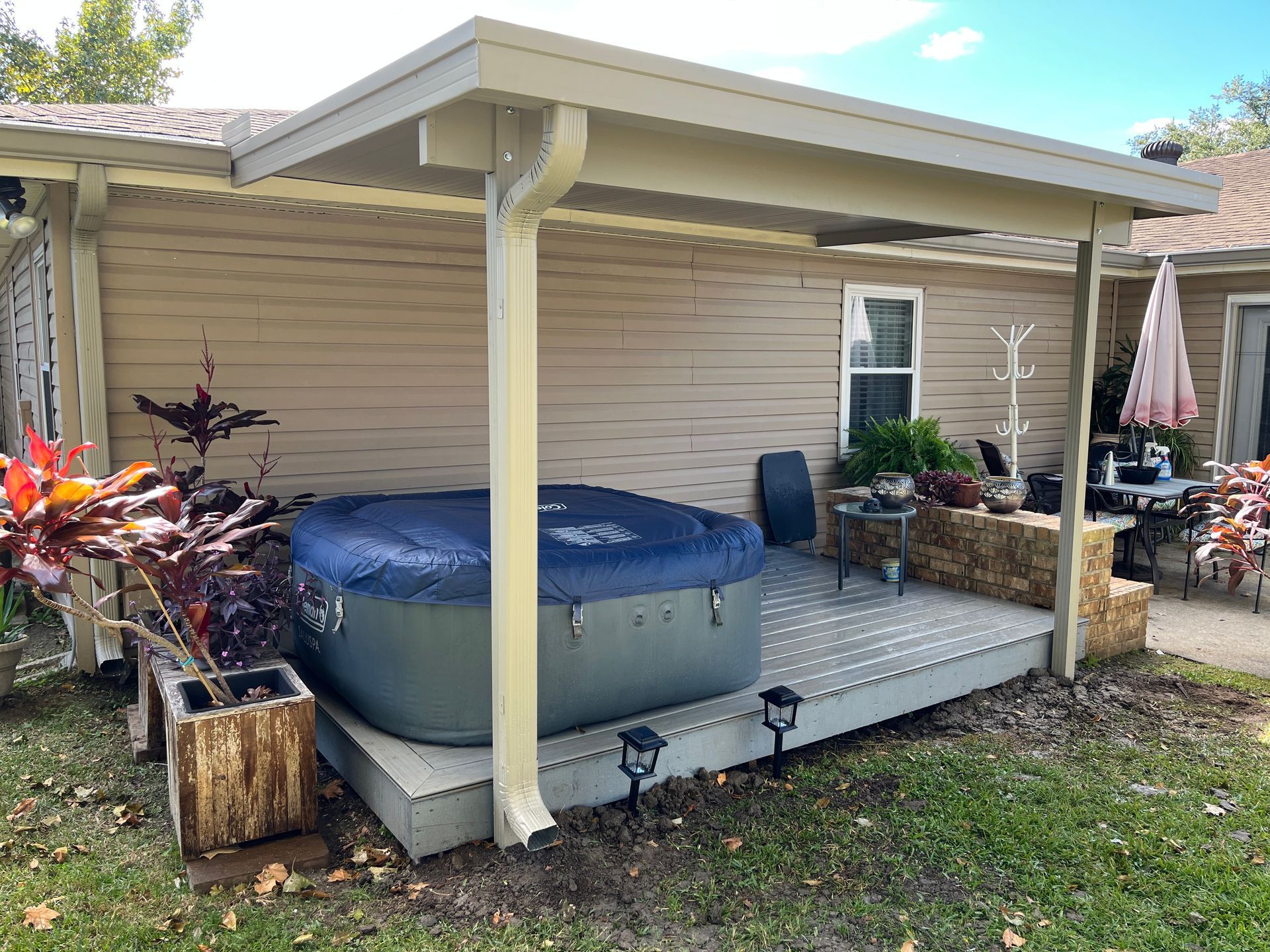 An outdoor deck covered by a patio roof features a blue-covered hot tub, a planter, and patio furniture.