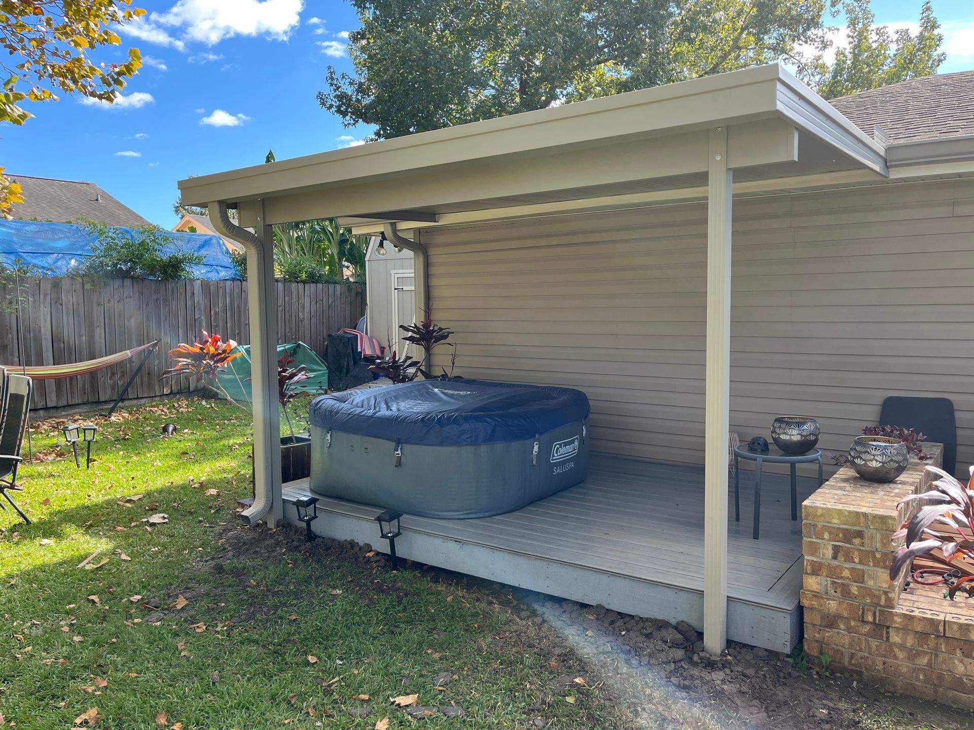 A covered patio with a gray hot tub, small table, and stone planter in a grassy backyard.