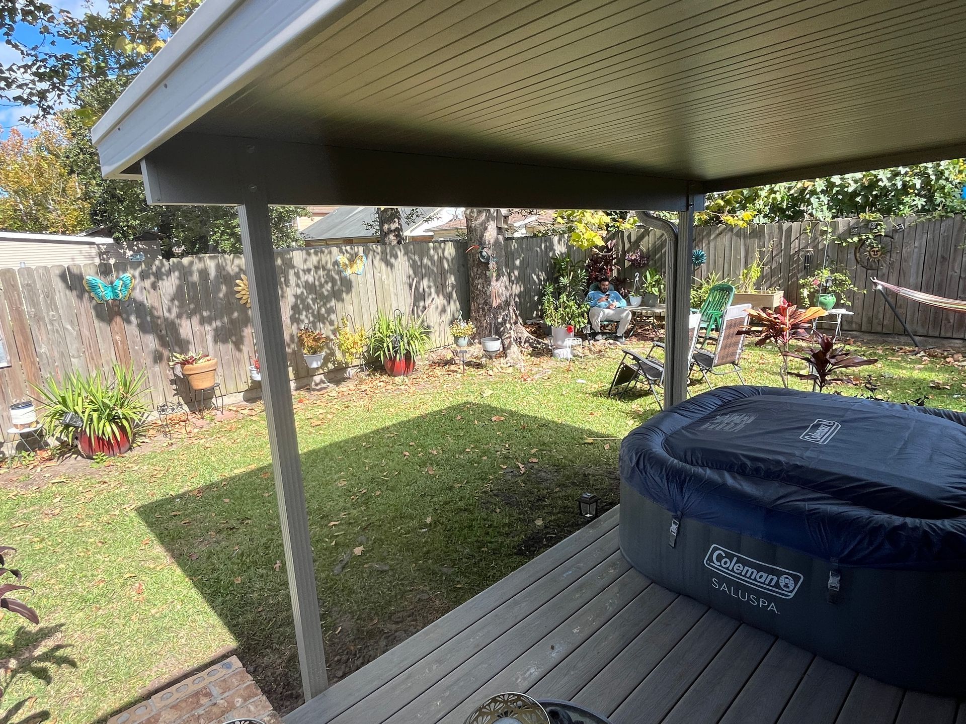 A covered wooden deck features an inflatable Coleman hot tub, overlooking a grassy backyard with a fence and garden.