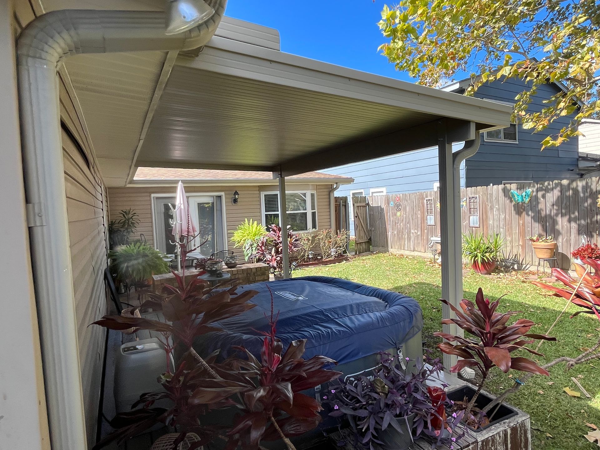 A hot tub with a blue cover sits under a patio roof in a backyard, surrounded by potted plants and a wooden fence.