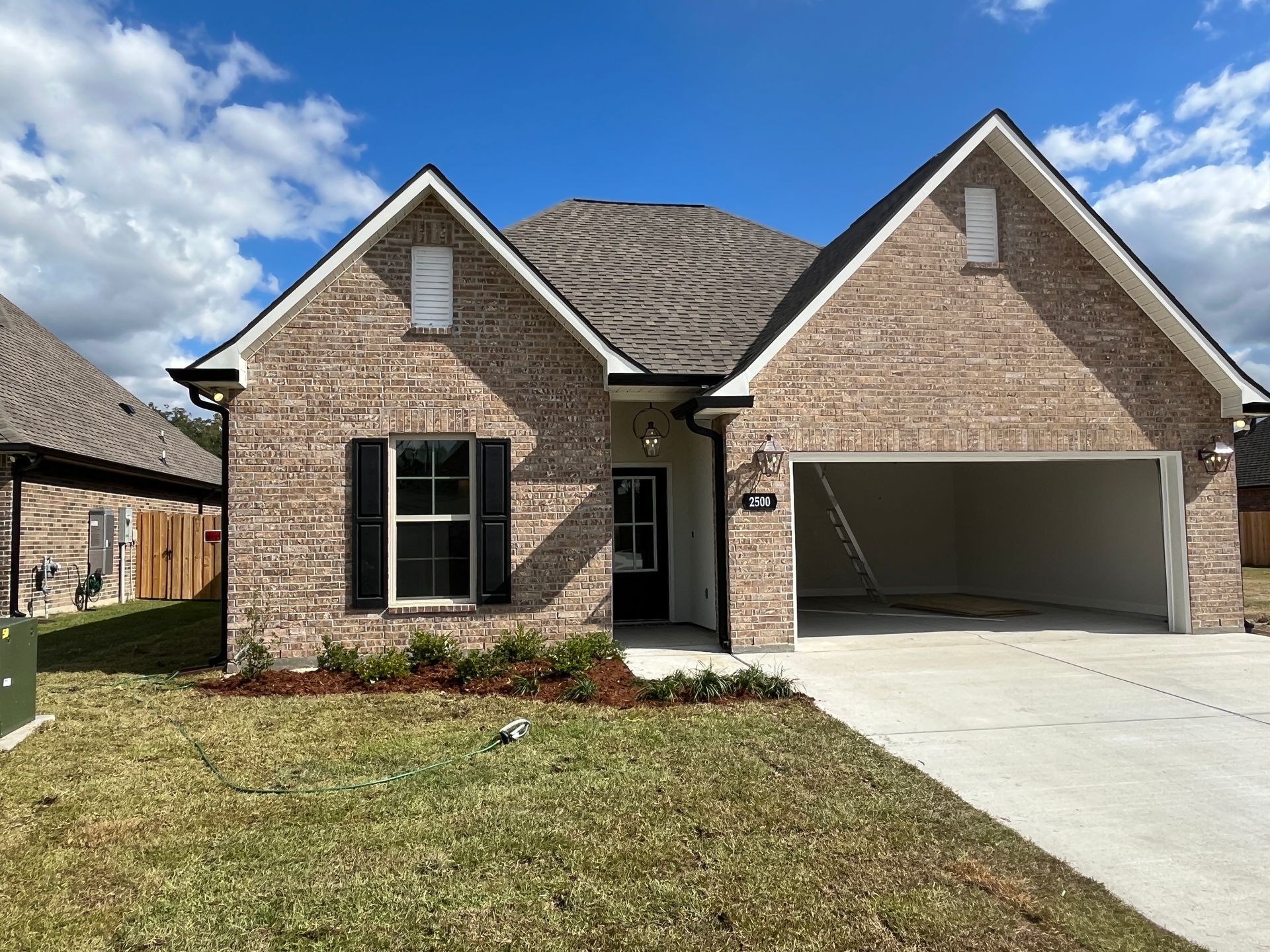 A one-story brick house with a two-car garage, black window shutters, and a front-facing gable under a bright blue sky.