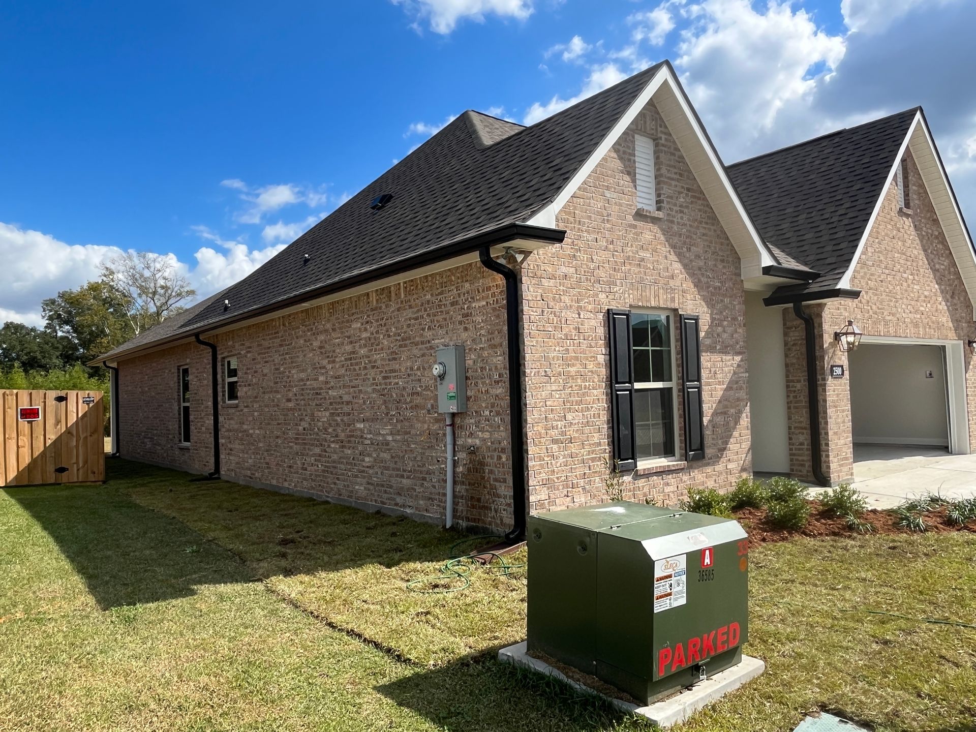 A tan brick house with a dark roof and a green electrical transformer box in the grassy front yard.